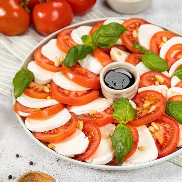 Skiver av mozzarella og tomat lagt i sirkel på et hvitt fat, pyntet med frisk basilikum og pinjekjerner, med en liten skål balsamicoeddik i midten. Hele tomater ligger i bakgrunnen.