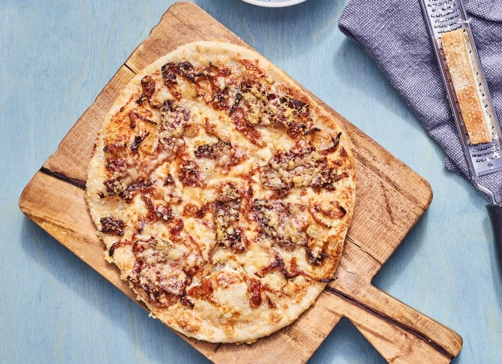 Tynn pizza med gyllen ost og karamellisert løk, servert på et rustikt skjærebrett av tre på et lyseblått bord.