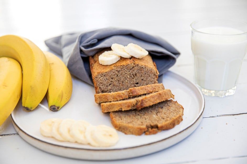 Hjemmelaget bananbrød på et fat, med flere skiver skåret opp, dekorert med bananskiver, friske bananer ved siden av og et glass melk i bakgrunnen.