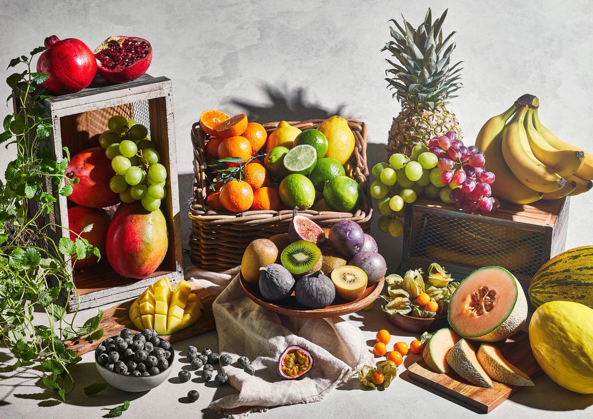 Et fargerikt stilleben med et bredt utvalg frukt arrangert på bord og i kasser, inkludert ananas, bananer, druer, appelsiner, sitrusfrukt, kiwi, granatepler, blåbær, fiken og melon, vist både hele og oppskåret i mykt, naturlig lys.