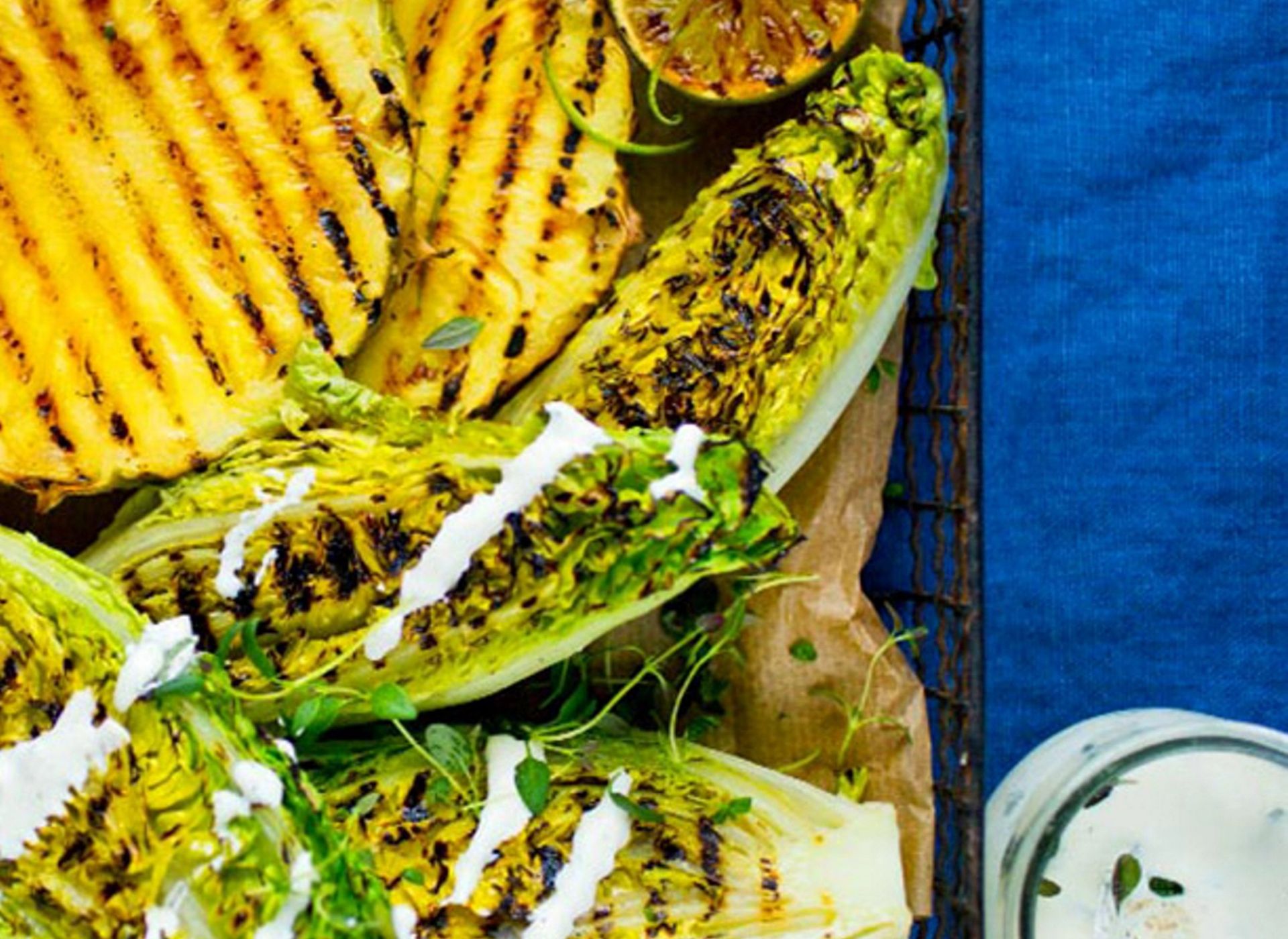 Grillet hjertesalat og skiver av ananas ligger på et fat, med tydelige grillmerker, små grønne blader og en lys dressing dryppet over, ved siden av en sitrushalvdel.