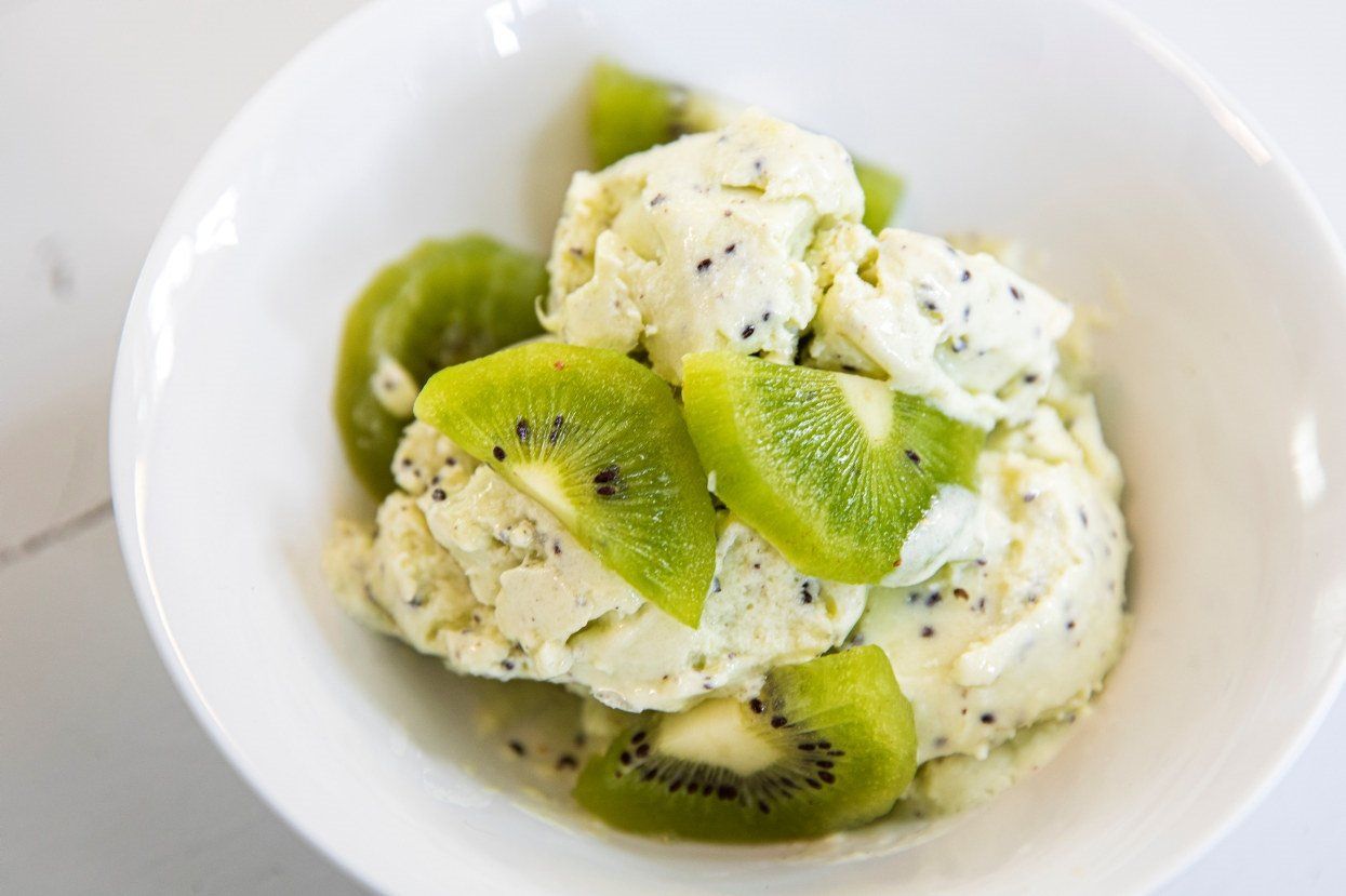 En hvit bolle fylt med kuler av lysegrønn kiwi-is, toppet med friske skiver av grønn kiwi. Isen er kremet med synlige små, svarte frø fra frukten.