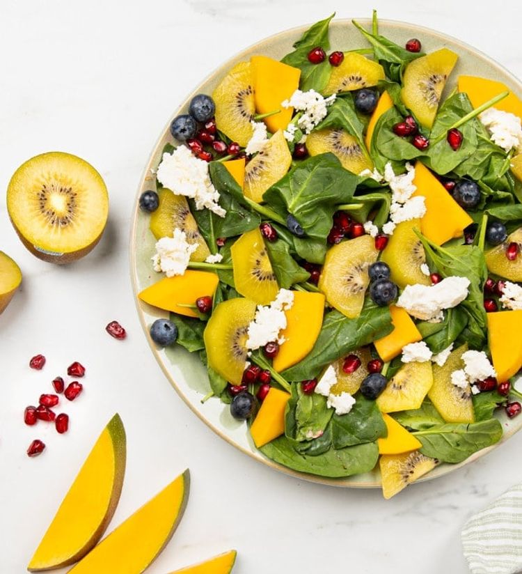 En fargerik salat anrettet på en lysegrønn tallerken mot en hvit marmorflate. Salaten består av frisk spinat, skiver av gul kiwi, biter av frisk mango, blåbær, granateplekjerner og smuldret fetaost.