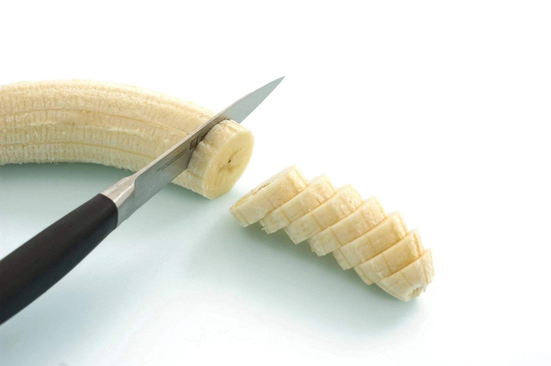 En skrelt banan som skjæres i skiver med en kniv på hvit bakgrunn.