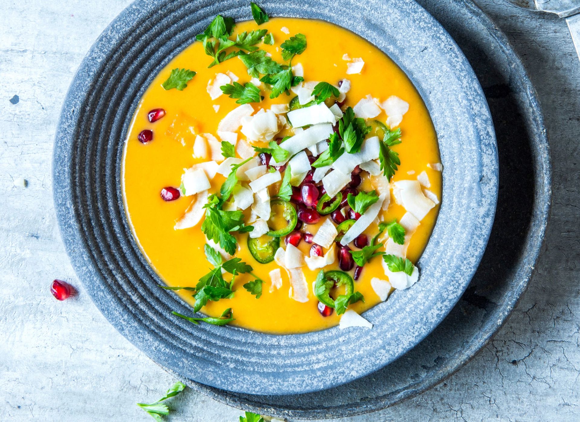 Kremet gresskarsuppe med søtpotet, rødløk, ingefær, lime og kokosmelk servert i en skål, toppet med skiver av chili, fersk koriander, granateplekjerner og kokosflak.