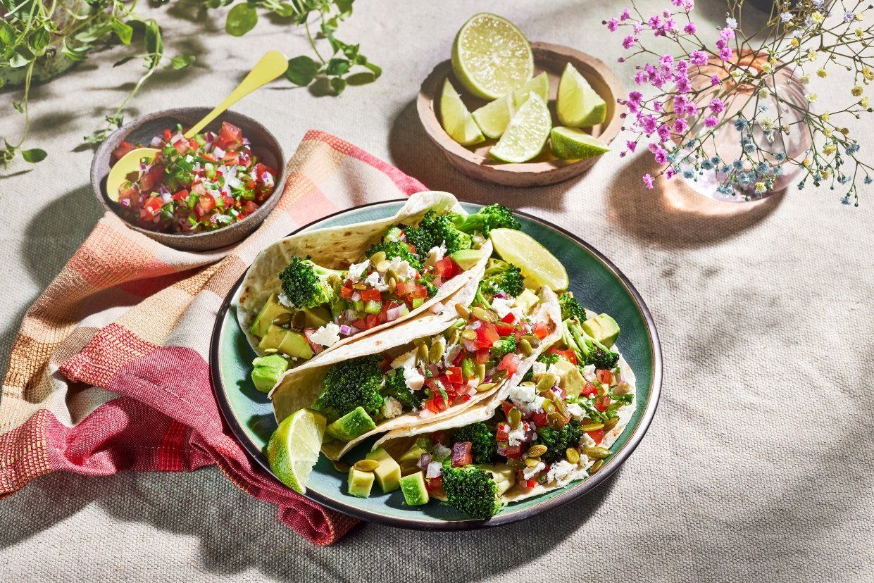 Tallerken med brokkolitacos fylt med avokado, tomatsalsa, fetaost og gresskarkjerner, servert på duk med ekstra salsa og lime i skåler ved siden av.