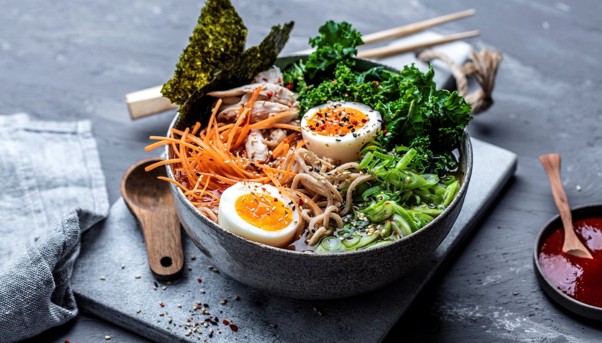 Kyllingramen servert i skål med nudler, saftig kylling, bløtkokt egg, grønnkål, vårløk, gulrot og nori på mørk bakgrunn.