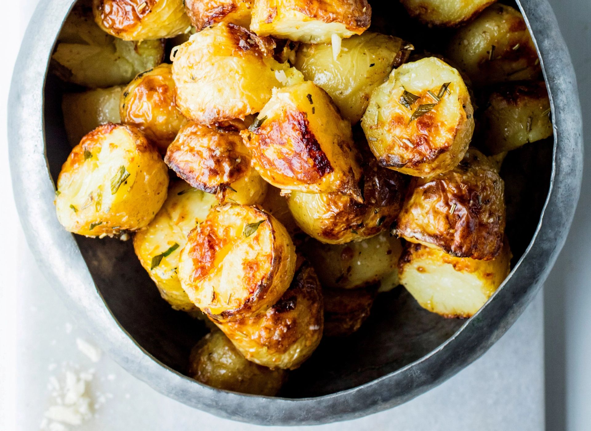 Ovnsbakte poteter med rosmarin og hvitløk, sprøstekt gylne og servert i en metallskål. Et dryss parmesan ligger på bordplaten ved siden.