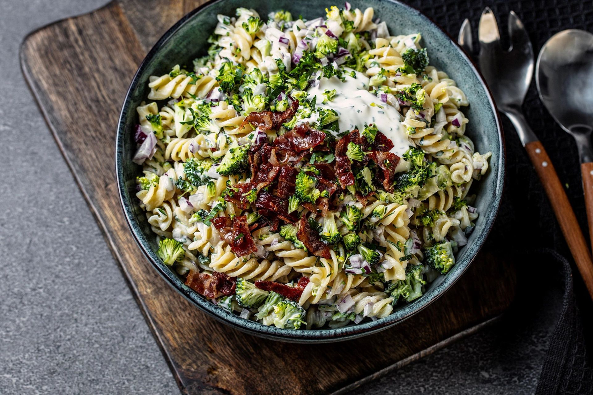 Skål med kremet pastasalat sett ovenfra, laget med fusilli, brokkoli, rødløk og sprø bacon, toppet med urtedressing. Skålen står på et rustikt treunderlag med bestikk ved siden av