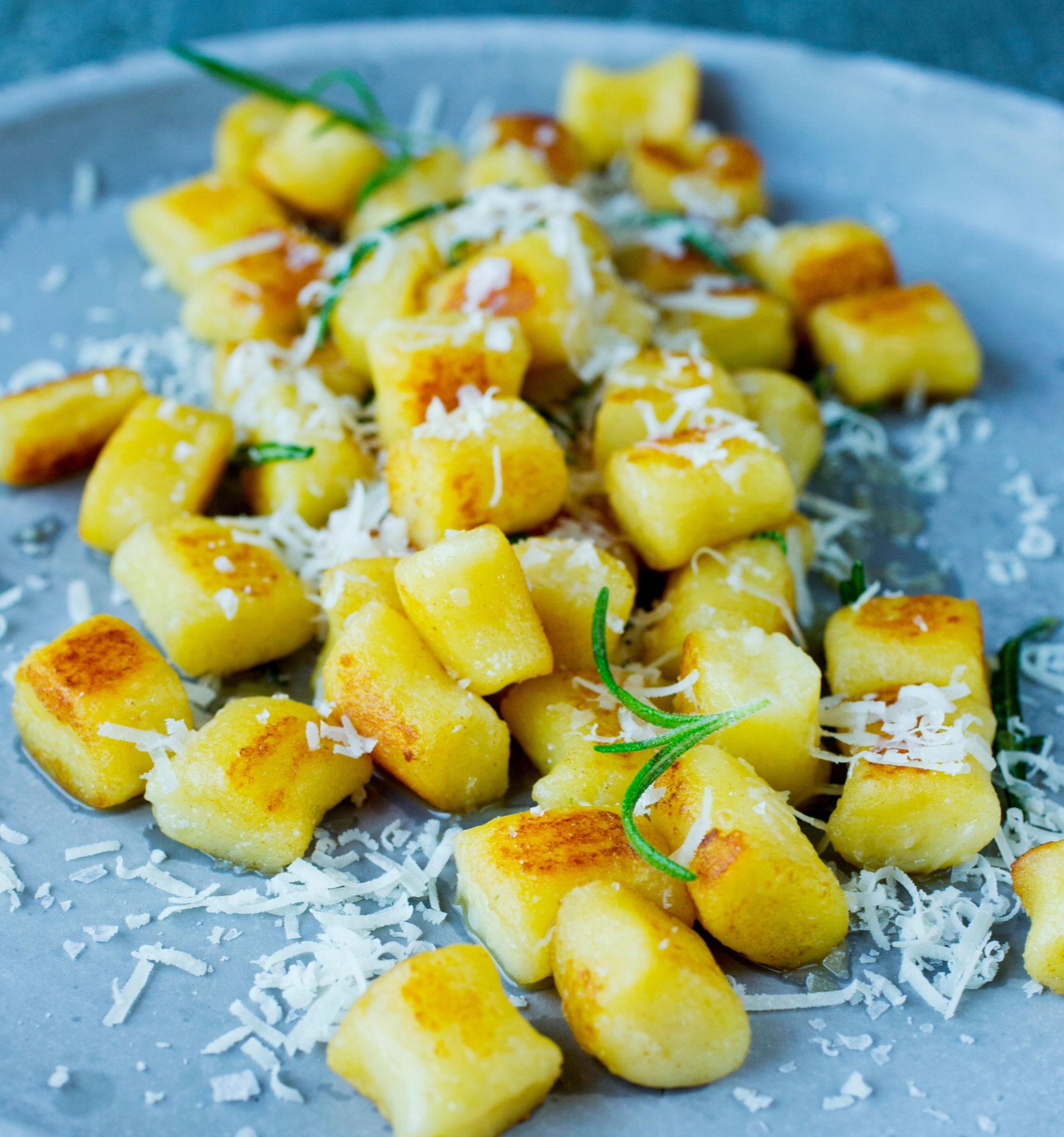 Stekte gnocchi‑biter med gyllen overflate servert på en grå tallerken, toppet med revet ost og små kvister frisk rosmarin.