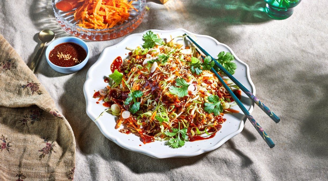 Spicy koreansk coleslaw laget av strimlet hodekål, gulrøtter og vårløk, toppet med koriander og sesamfrø, servert på et hvitt fat med spisepinner og tilbehør i bakgrunnen.
