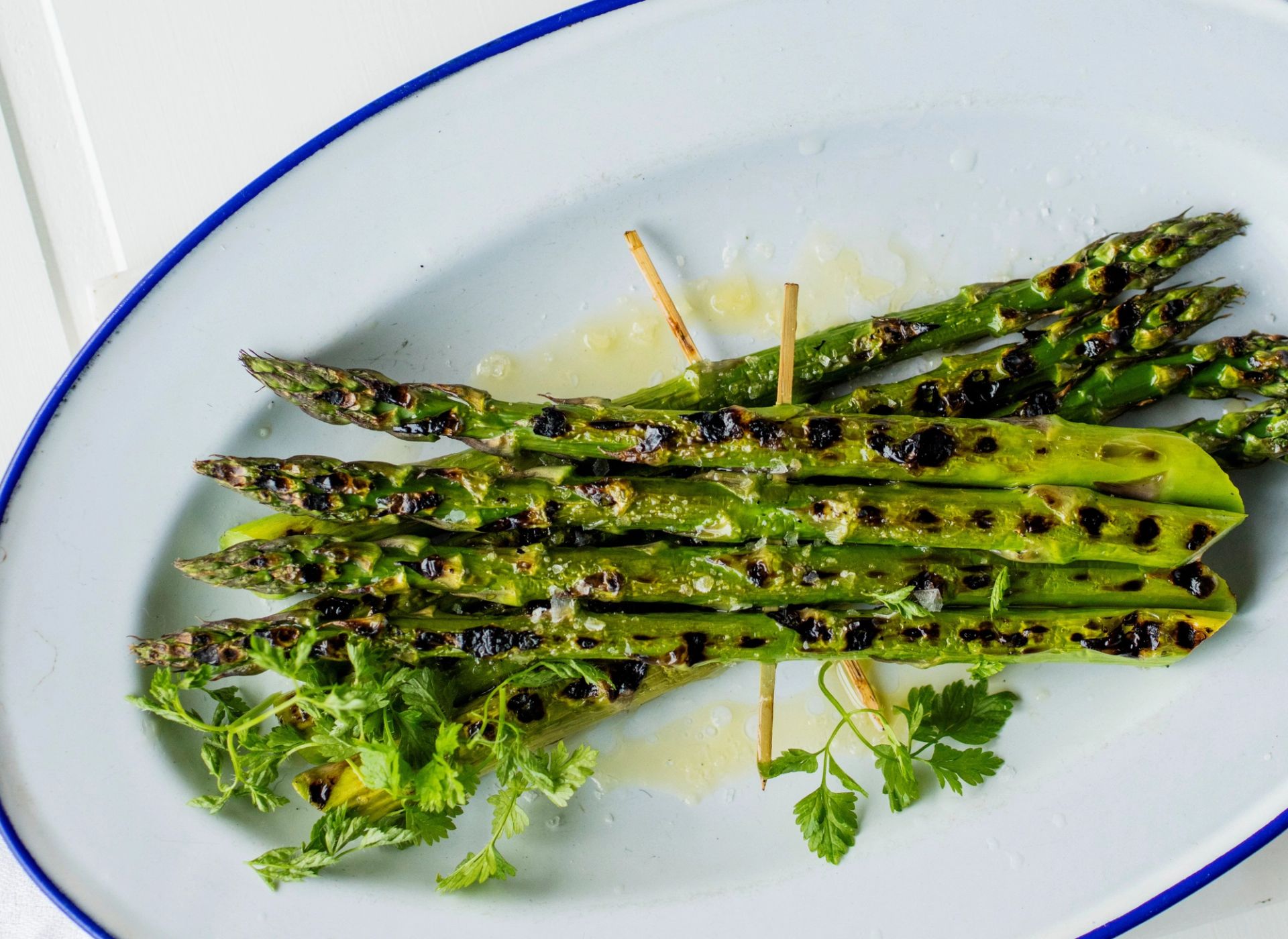 Grillet grønn asparges med olivenolje og sitron, servert på et lyst fat. De grillede aspargesene har tydelige grillmerker og er toppet med friske urter. Enkel og sunn grønnsaksrett med sitron og olivenolje.