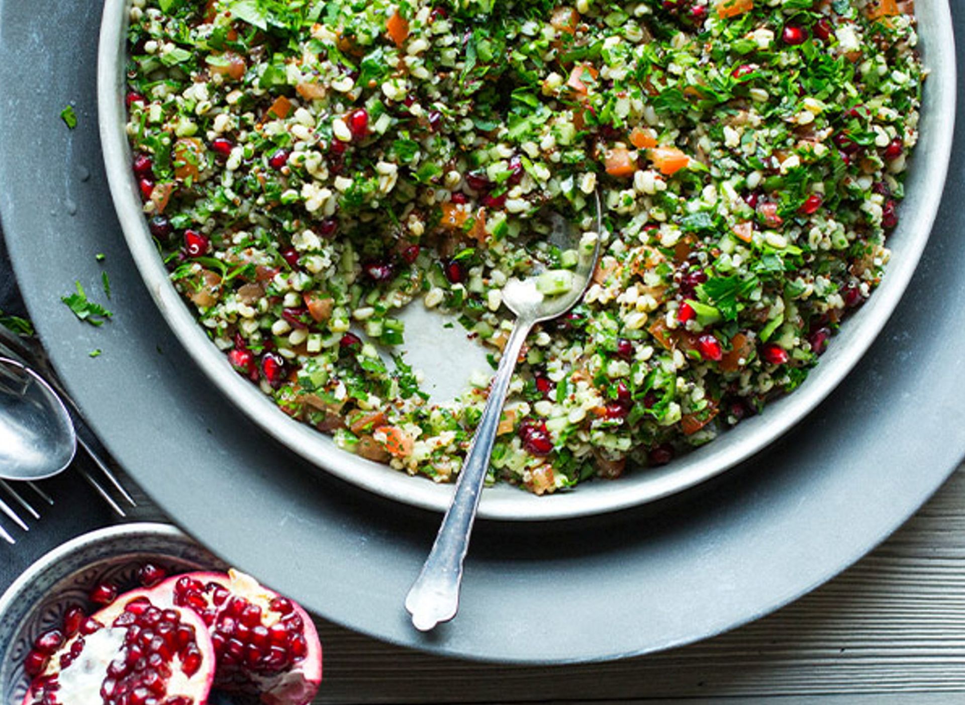 En stor bolle med finhakket salat med bulgur, grønne urter, små biter av grønnsaker og røde granateplekjerner. En skje ligger i bollen, og ved siden av står en delt granateplefrukt på et bord.