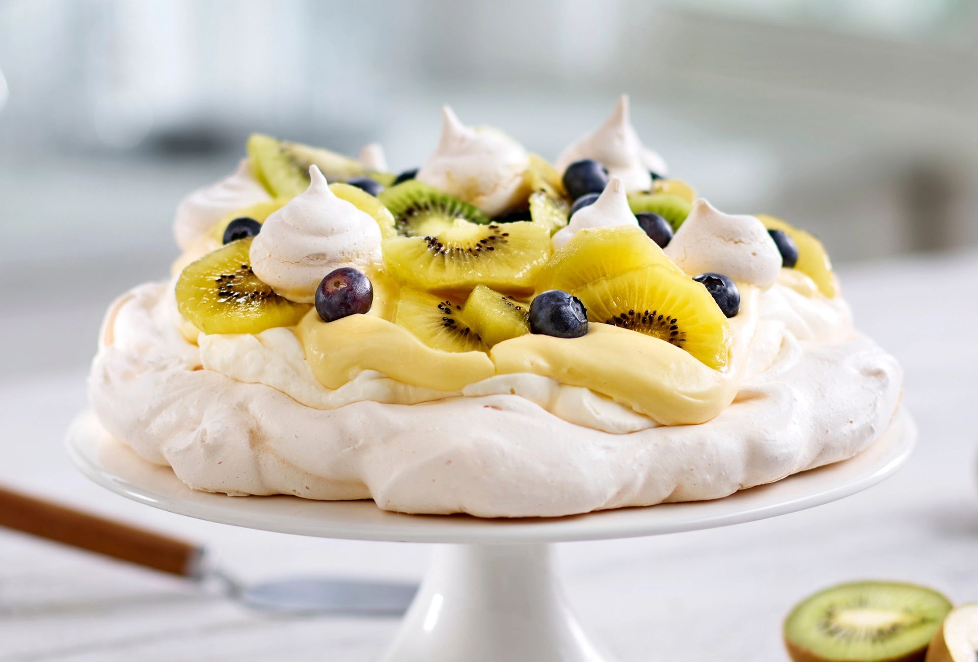 En pavlova står på et hvitt kakefat. Den består av en stor, lys marengsbunn toppet med et lag krem og gul vaniljekrem. På toppen ligger skiver av kiwi og hele blåbær, samt små marengstopper. I forgrunnen til høyre ligger en halv oppskåret kiwi. Bakgrunnen er lys og uklar, og gir et luftig, mykt uttrykk.
