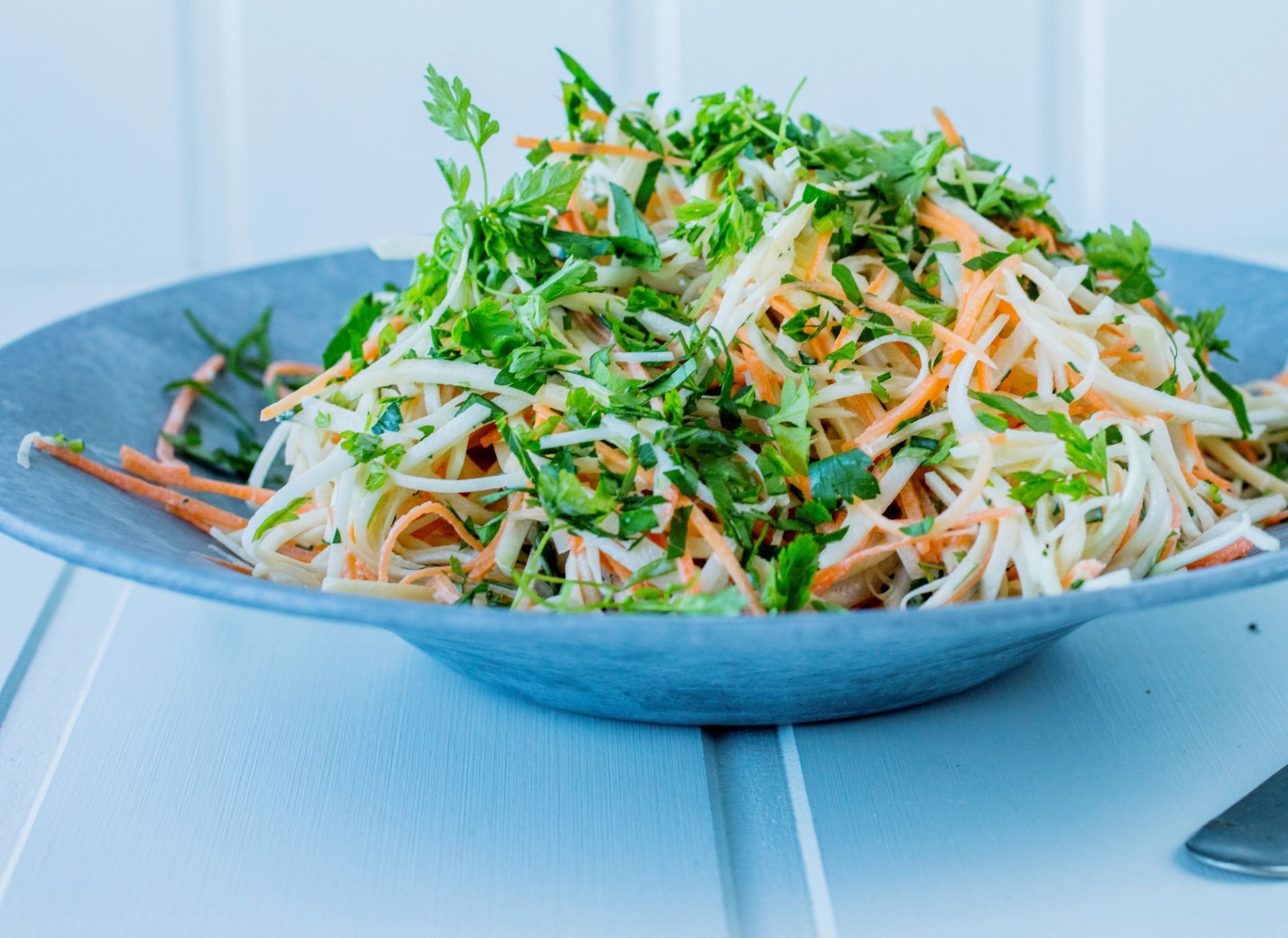 En stor porsjon hjemmelaget coleslaw servert i en bred, gråblå bolle. Salaten består av finsnittet hodekål og revne gulrøtter, toppet med en generøs mengde hakket frisk bladpersille. Bollen står på et lyst blåmalt tregulv.