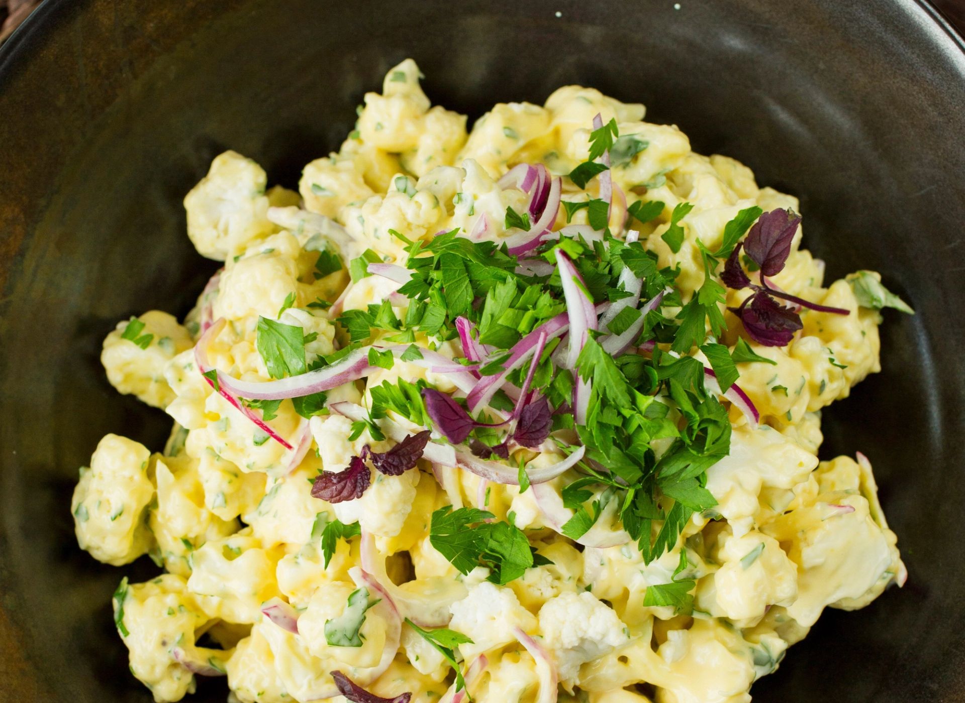 En kremet blomkålsalat blandet med majonesdressing, servert i en mørk bolle og toppet med grovhakket persille og tynne skiver rødløk.
