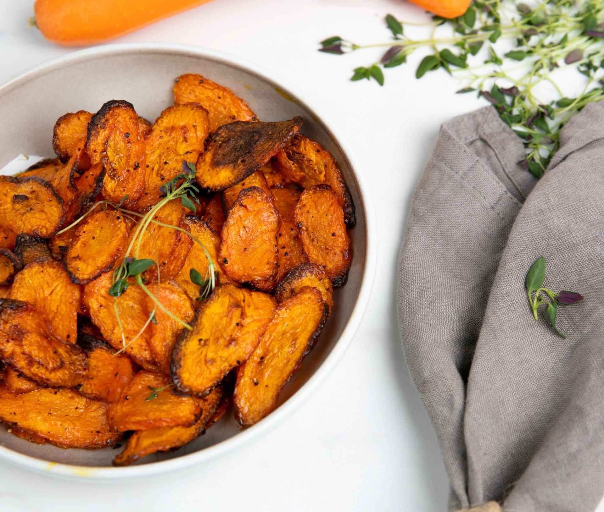 Sprøstekte airfryer gulrotchips i en beige bolle, krydret med timian og pepper. Friske urter, hele gulrøtter og en grå kjøkkenklut ligger rundt på en hvit benkeplate. Sunn og enkel snack laget i airfryer.