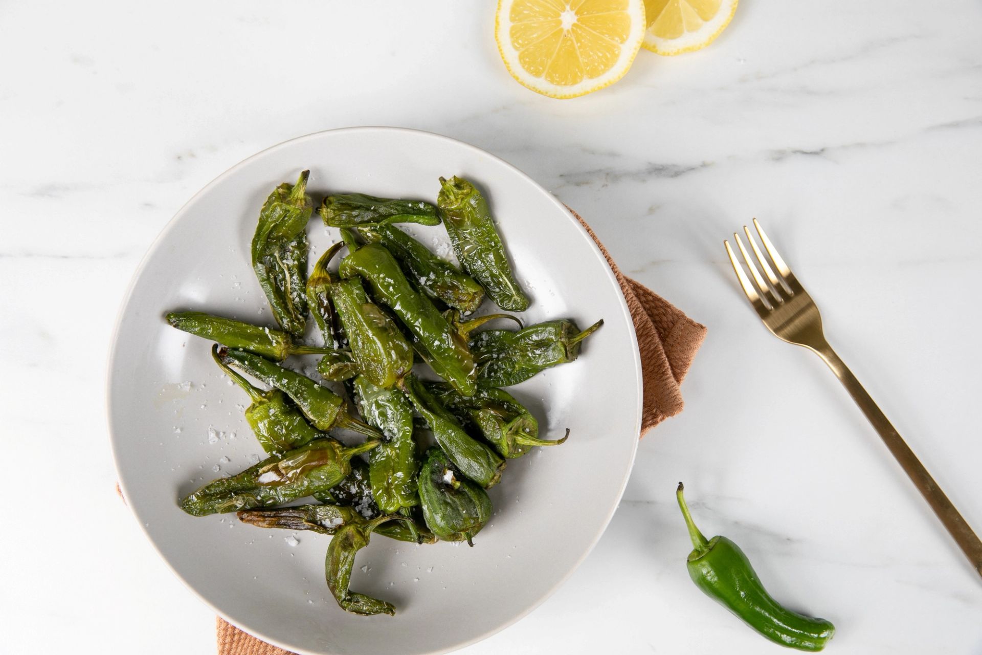 Sprø padron-paprika servert på en lys tallerken, toppet med flaksalt. Sitronskiver, en hel grønn padron og en gullfarget gaffel ligger ved siden av på en hvit benkeplate. Enkel og smakfull tapas laget i airfryer.