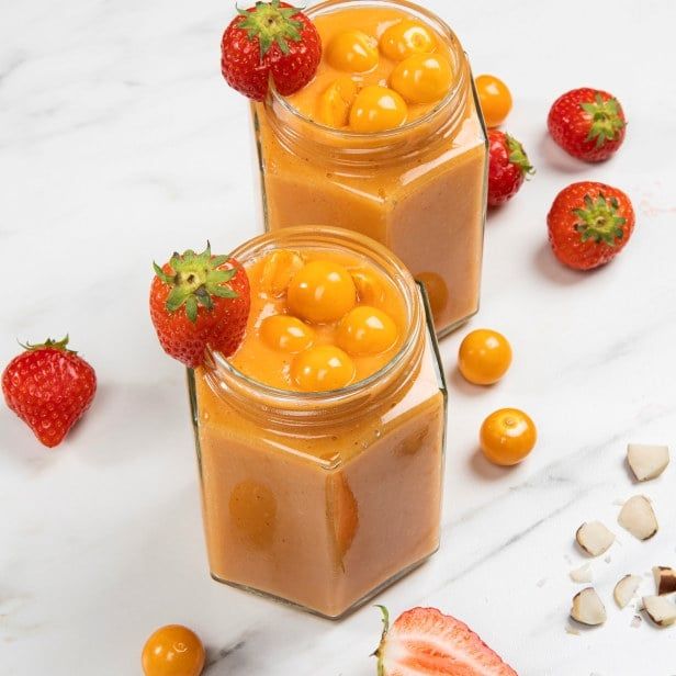 To sekskantede glass fylt med tykk, oransje mango- og physalissmoothie toppet med runde, gule physalisbær. Hvert glass er pyntet med et friskt jordbær på kanten. Rundt glassene ligger hele jordbær, physalisbær og noen små biter av kokos på en lys marmorlignende overflate.