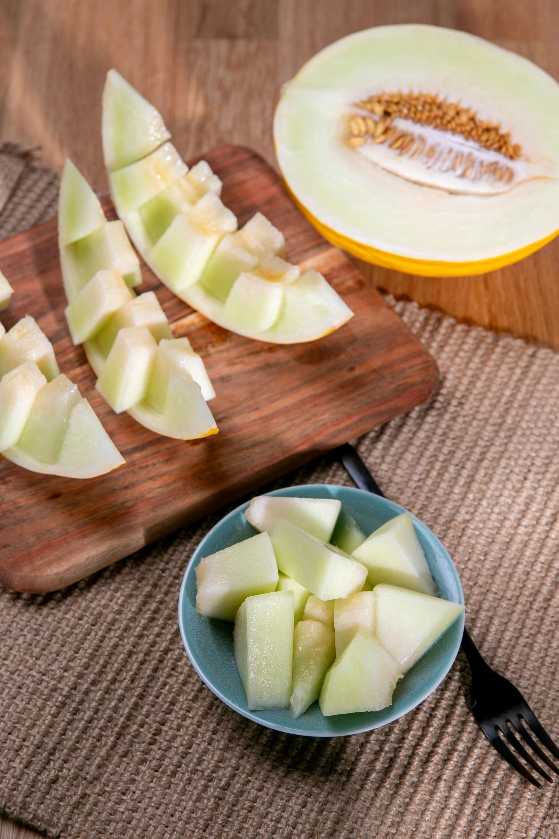 En halv honningmelon ligger i bakgrunnen, mens skiver og terninger av lys grønn melon er lagt på et skjærebrett og i en liten blå bolle.