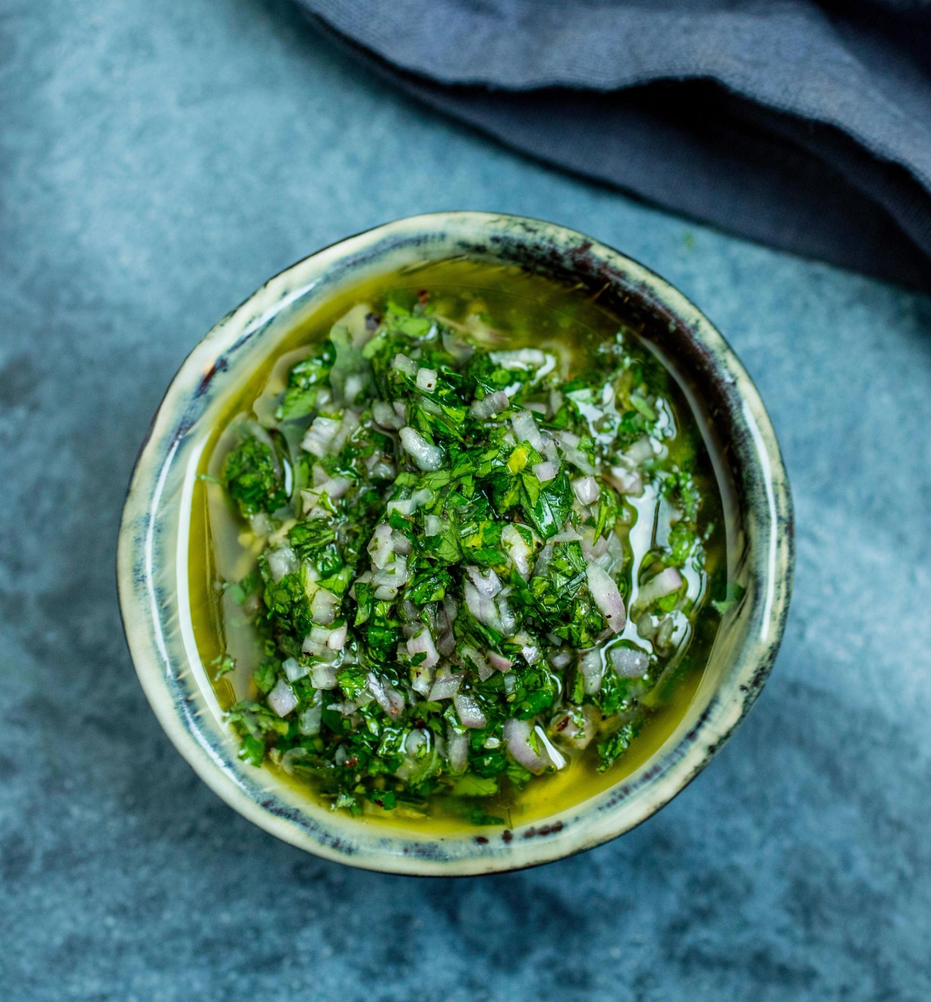 En liten bolle med hjemmelaget grønn saus laget av mynte og finhakkede urter i olje, lik en chimichurri, plassert på en blågrå overflate