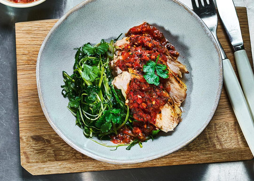 Tallerken med skiver av lyst kjøtt toppet med en grov tomat- og chilisaus, servert med sautert grønnkål. Retten står på et trebord på en stålbenk, omgitt av ferske tomater, urter og bestikk.