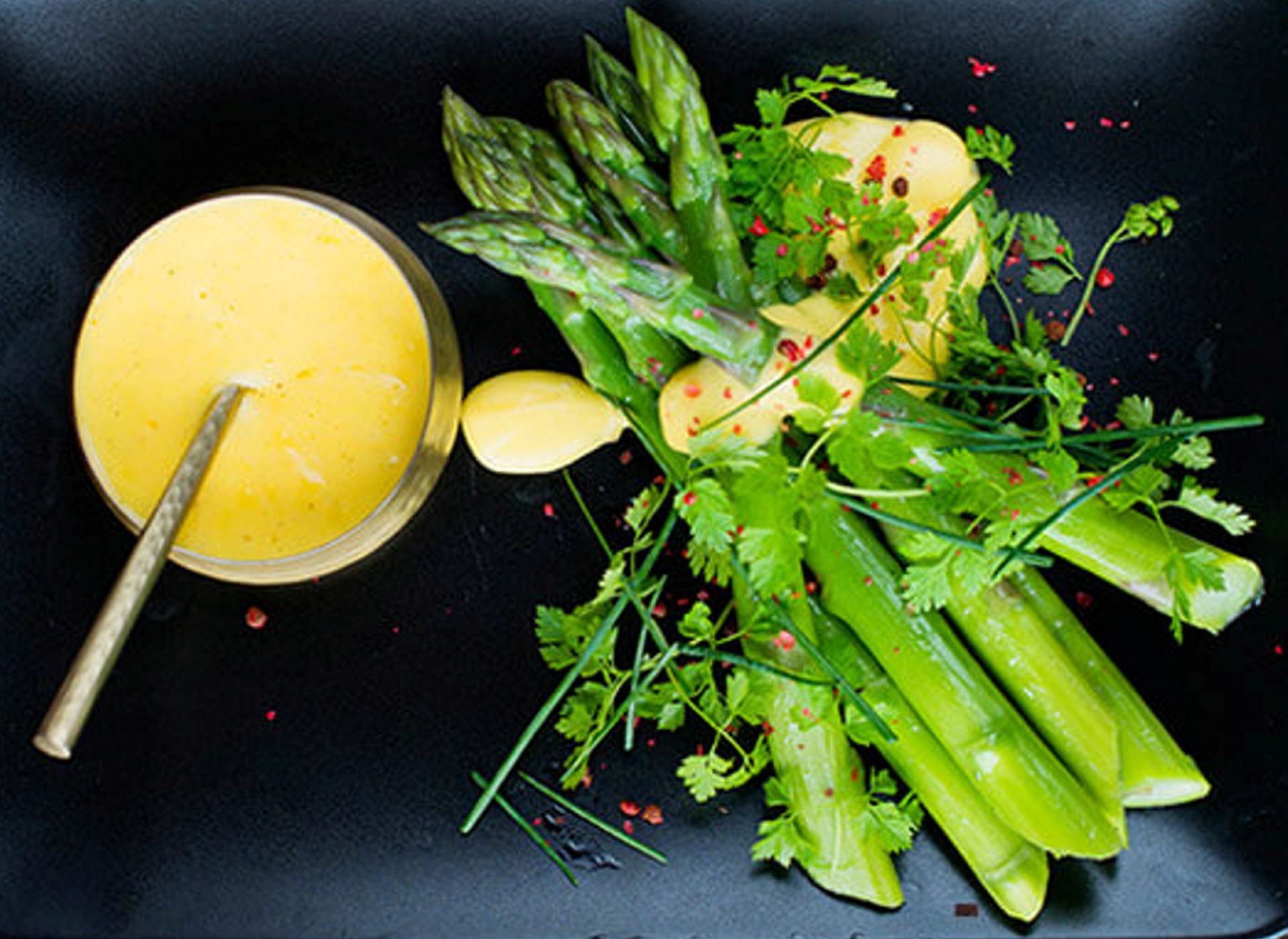 En sort tallerken med en liten glassbolle fylt med lys gul saus og en skje. Ved siden av ligger grønne aspargesstengler pyntet med friske urter og noen små rødrosa pepperkorn. Retten er dekorert med en lys, kremet saus og ser delikat anrettet ut.
