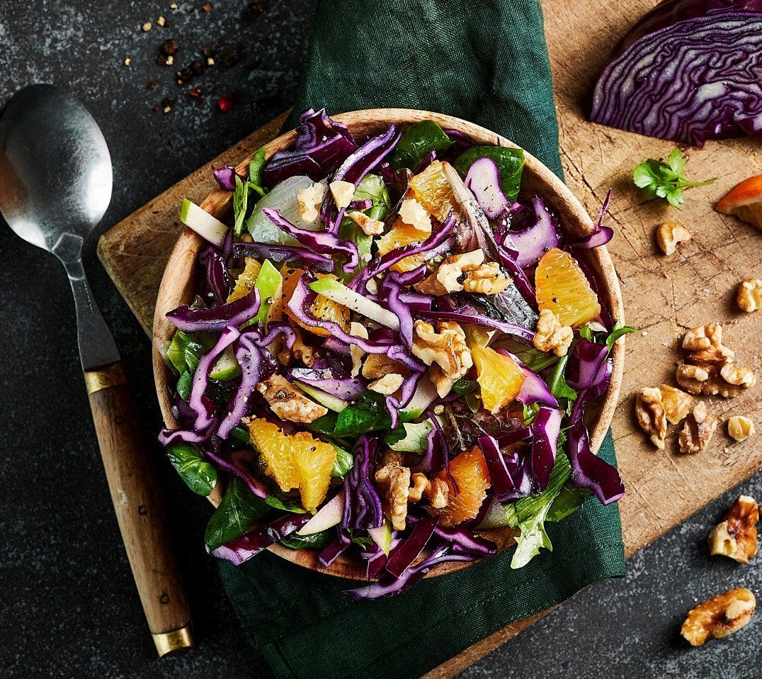 Salat av rødkål, appelsinbåter, epleskiver, salat og valnøtter ligger i en treskål, på en fjøl pyntet med valnøtter og en rødkålkile.