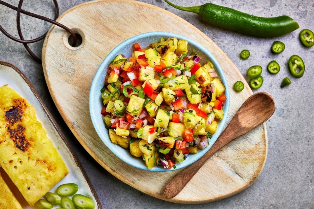 Skål med frisk salsa av grillede ananasbiter, rød paprika, rødløk, grønne urter og skivet chili, servert på et trefat med treskje.
