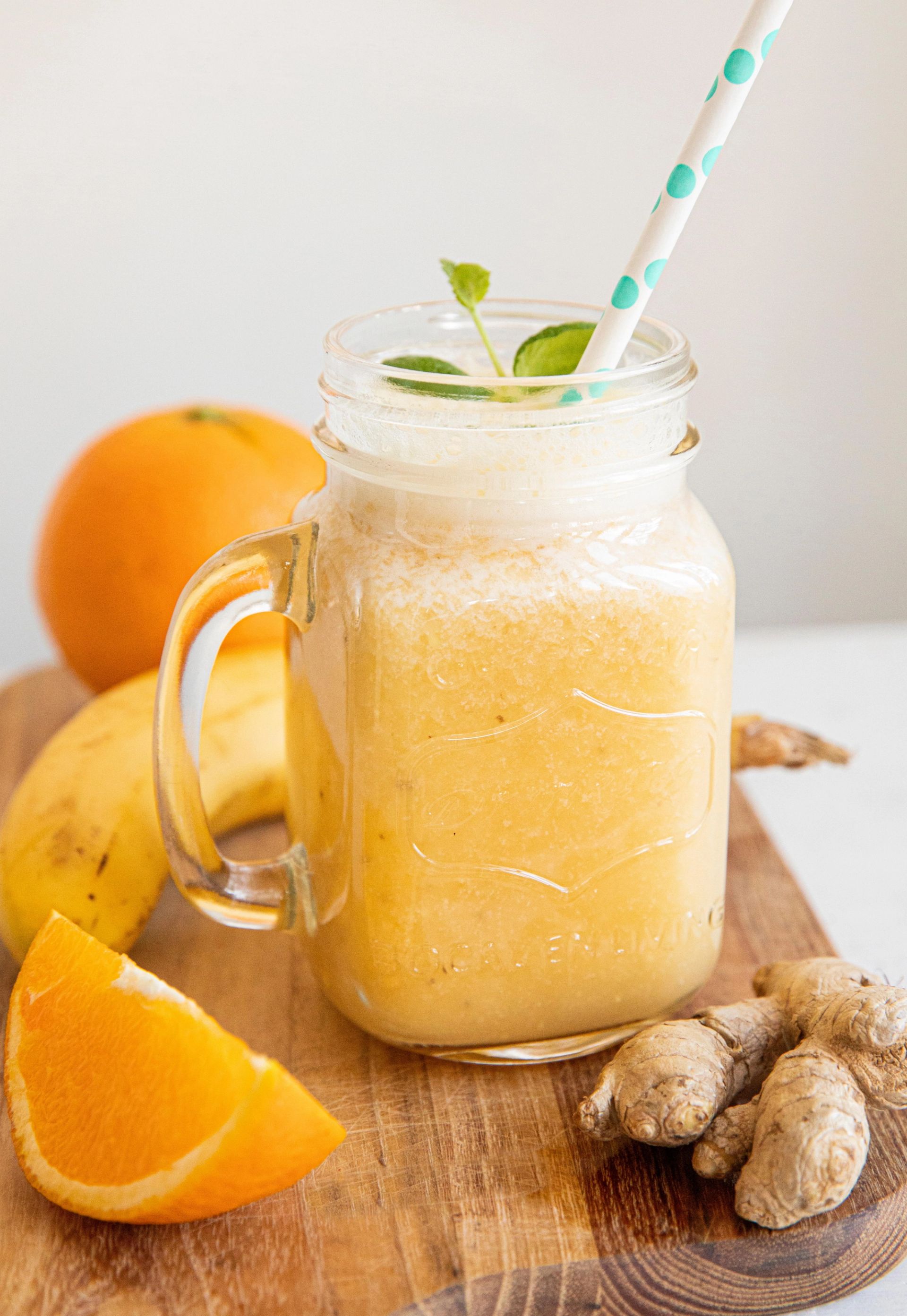 Glasskrukke med lys gul smoothie laget av appelsin, banan og ingefær, pyntet med sugerør og mynteblader, omgitt av fersk frukt og ingefær på en trefjøl