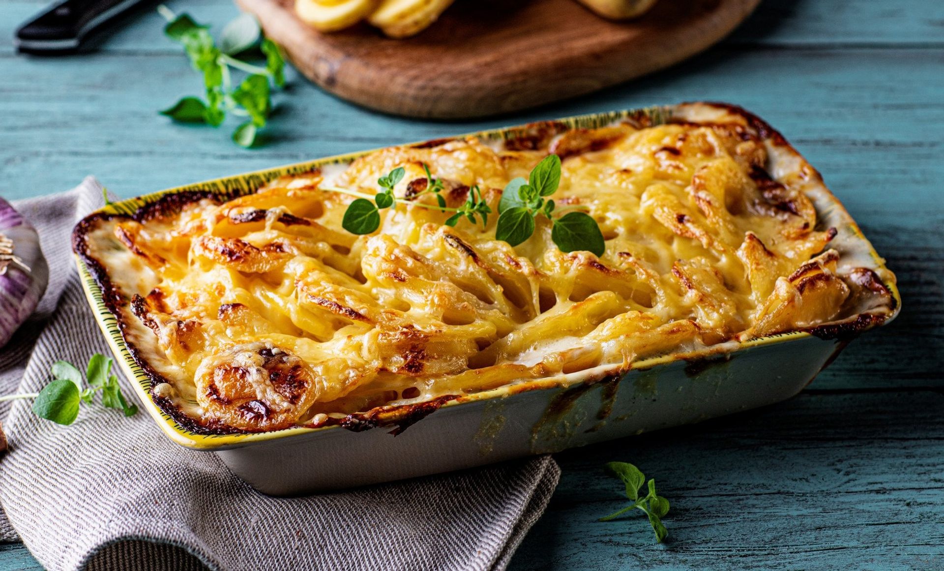 En gratengform fylt med gyllen, gratinert fløtegratinerte poteter, toppet med smeltet ost og friske urter. Formen står på et blått trebord med et kjøkkenhåndkle under.