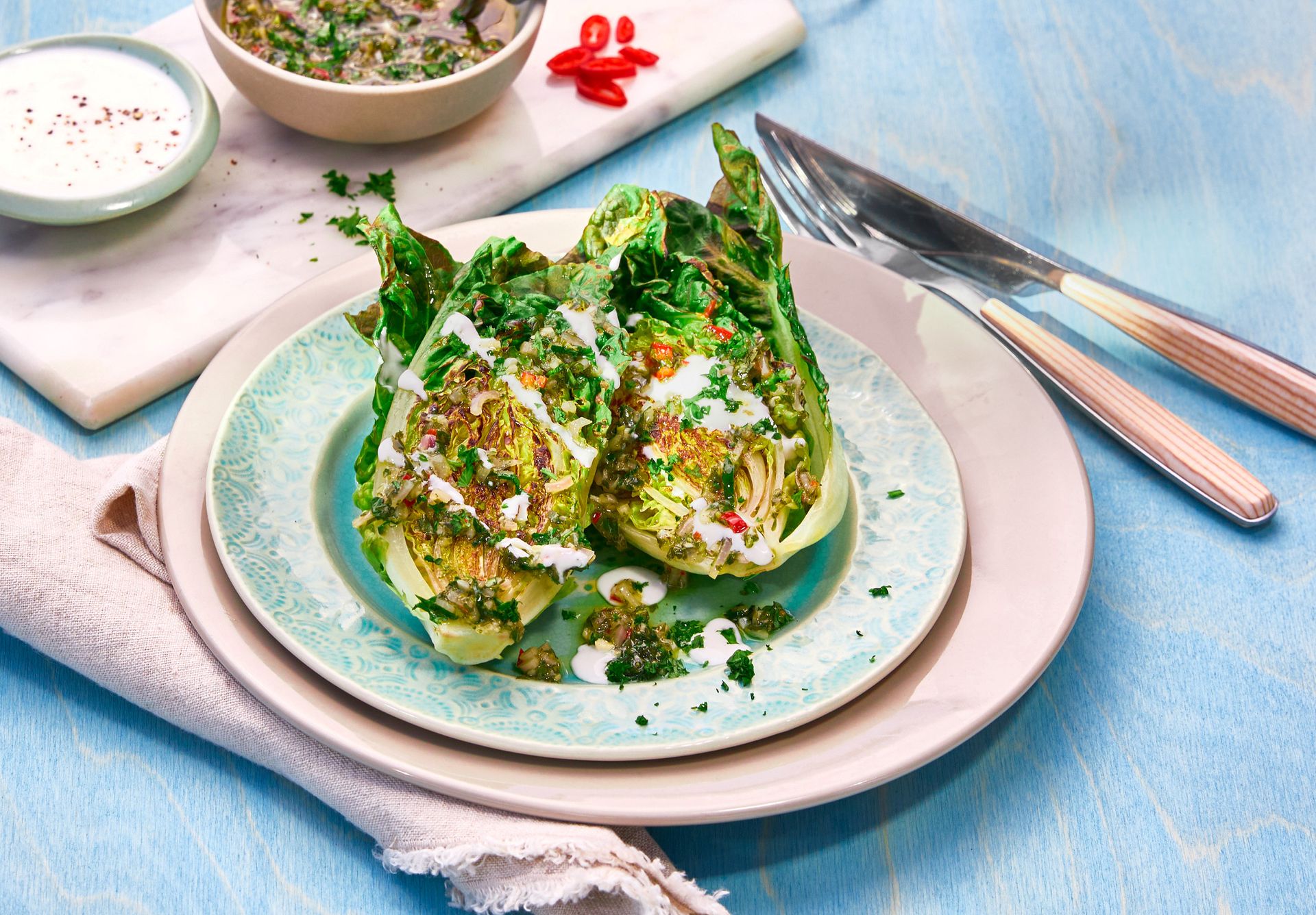 Oversiktsbilde av to grillede halve hjertesalat servert på en mønstret tallerken med lyseblå og rosa kanter. Salatene er generøst toppet med grønn chimichurri-saus og dryppet med hvit yoghurtdressing, pyntet med finhakkede urter og biter av rød chili.