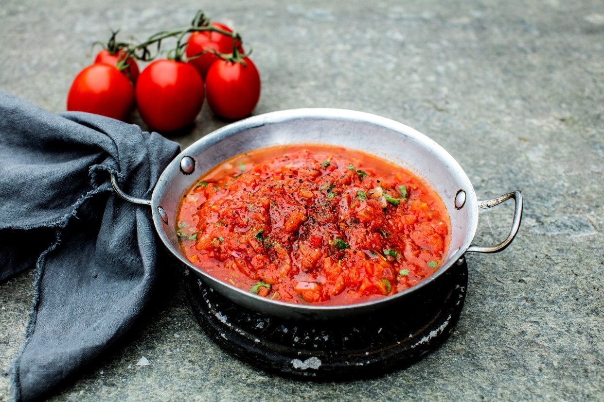En lav, rund metallpanne fylt med tomatbasert saus med synlige tomatbiter og urter står på et steinunderlag; ved siden av ligger en mørk kjøkkenklut.