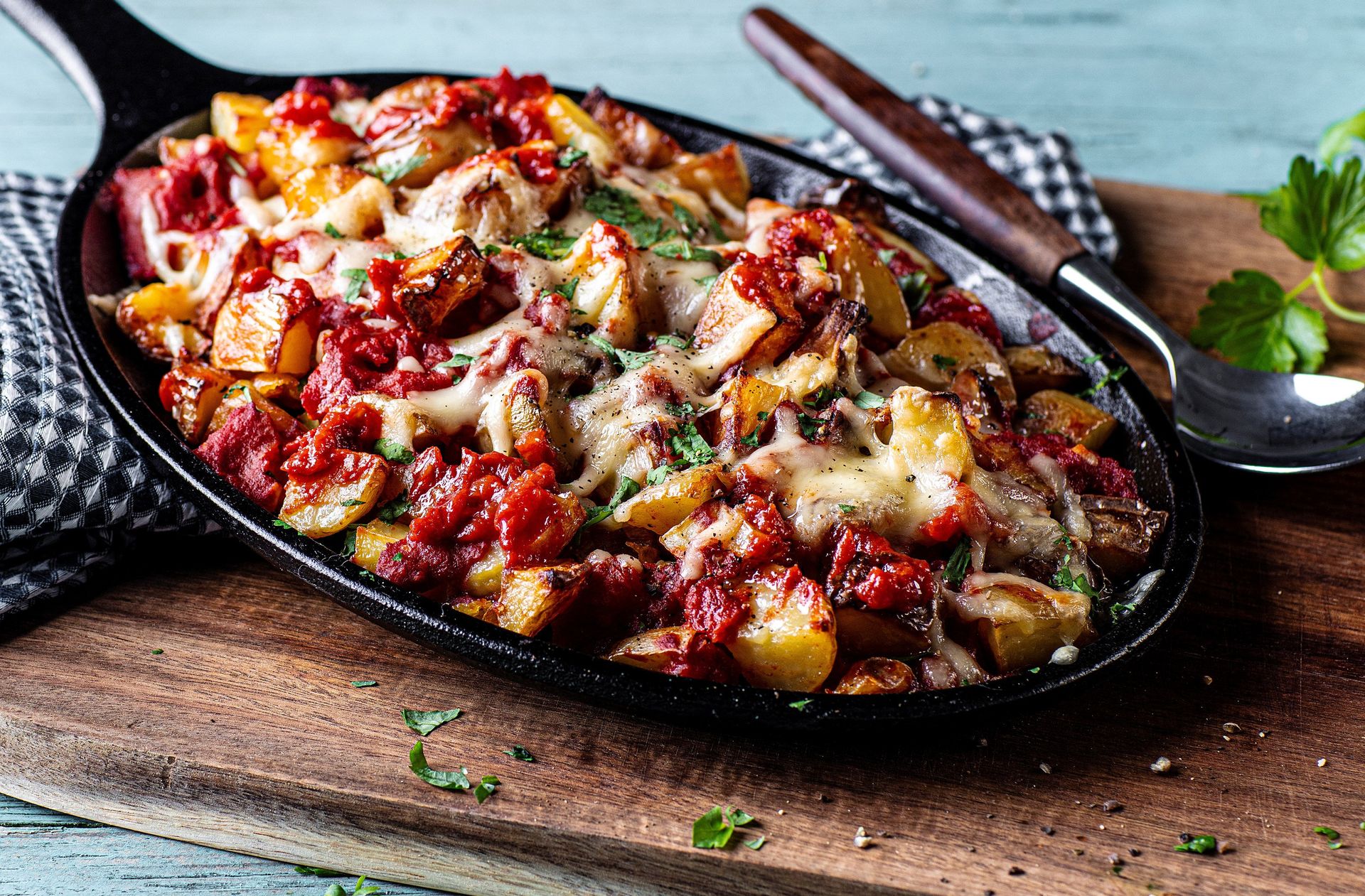 Et nærbilde av en varm stekepanne fylt med ovnsbakte poteter, toppet med smeltet ost og en chunky tomatsaus. Retten er drysset med friske urter og ser gyllen og sprø ut, med et serveringsbestikk ved siden av på et trebord.