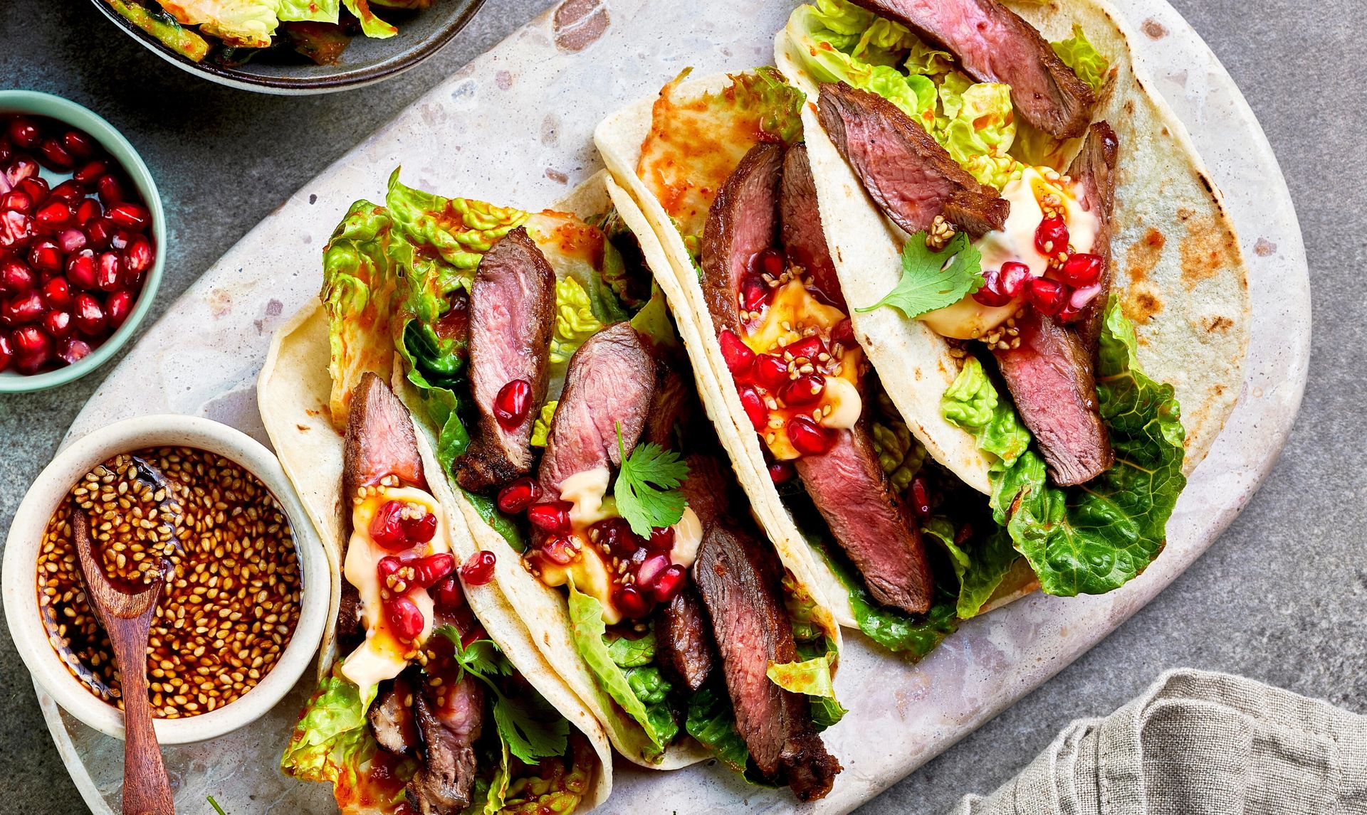 Tacos med marinert oksekjøtt, kimchi, sprø salat og granateplekjerner toppet med sesam dressing på lyst bord.