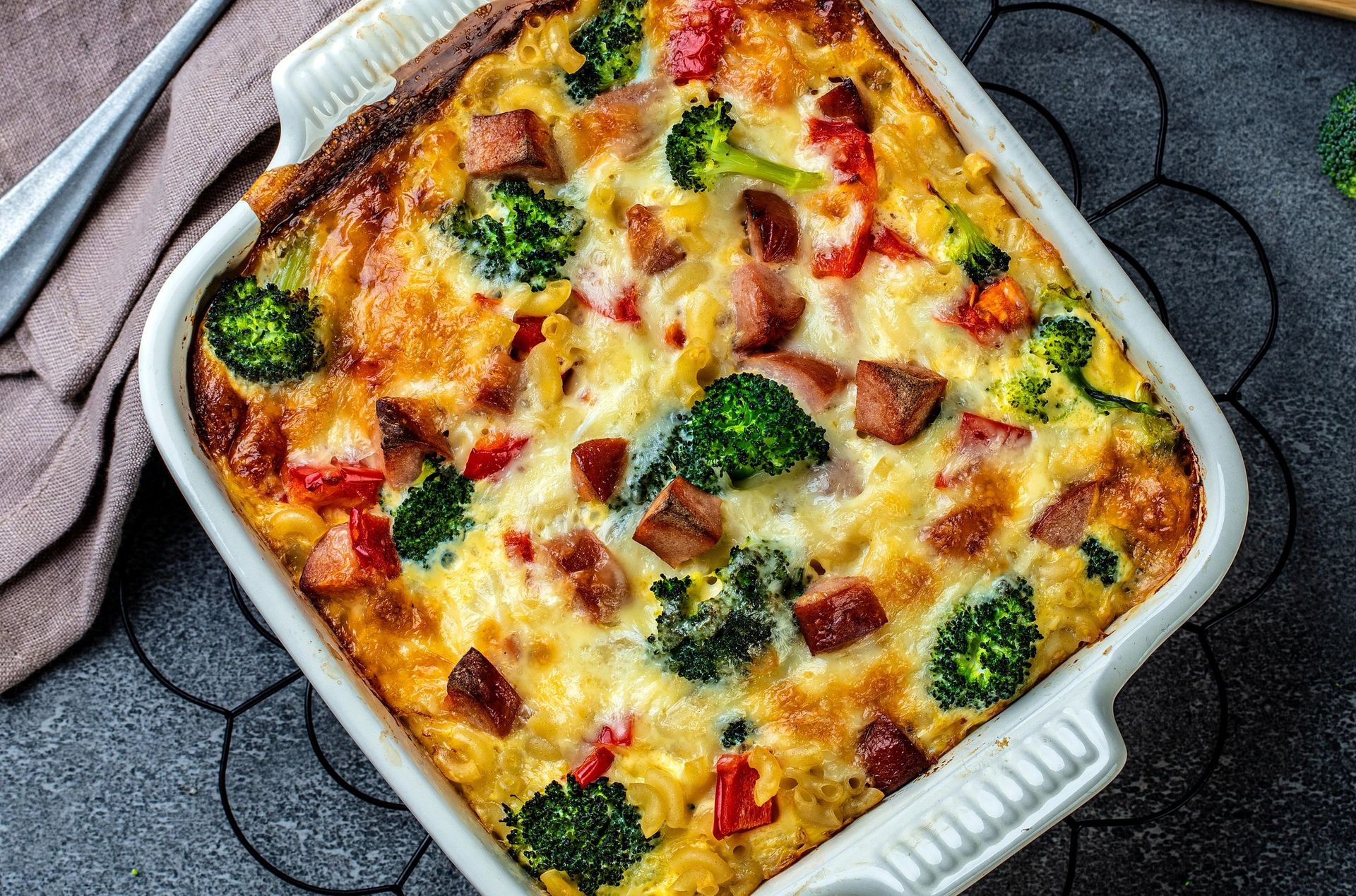 Ovnsbakt makaroniform med brokkoli, skinke og smeltet ost i en ildfast form, omgitt av ferske brokkolibuketter, en treskjærefjøl og en skje på et grått underlag.