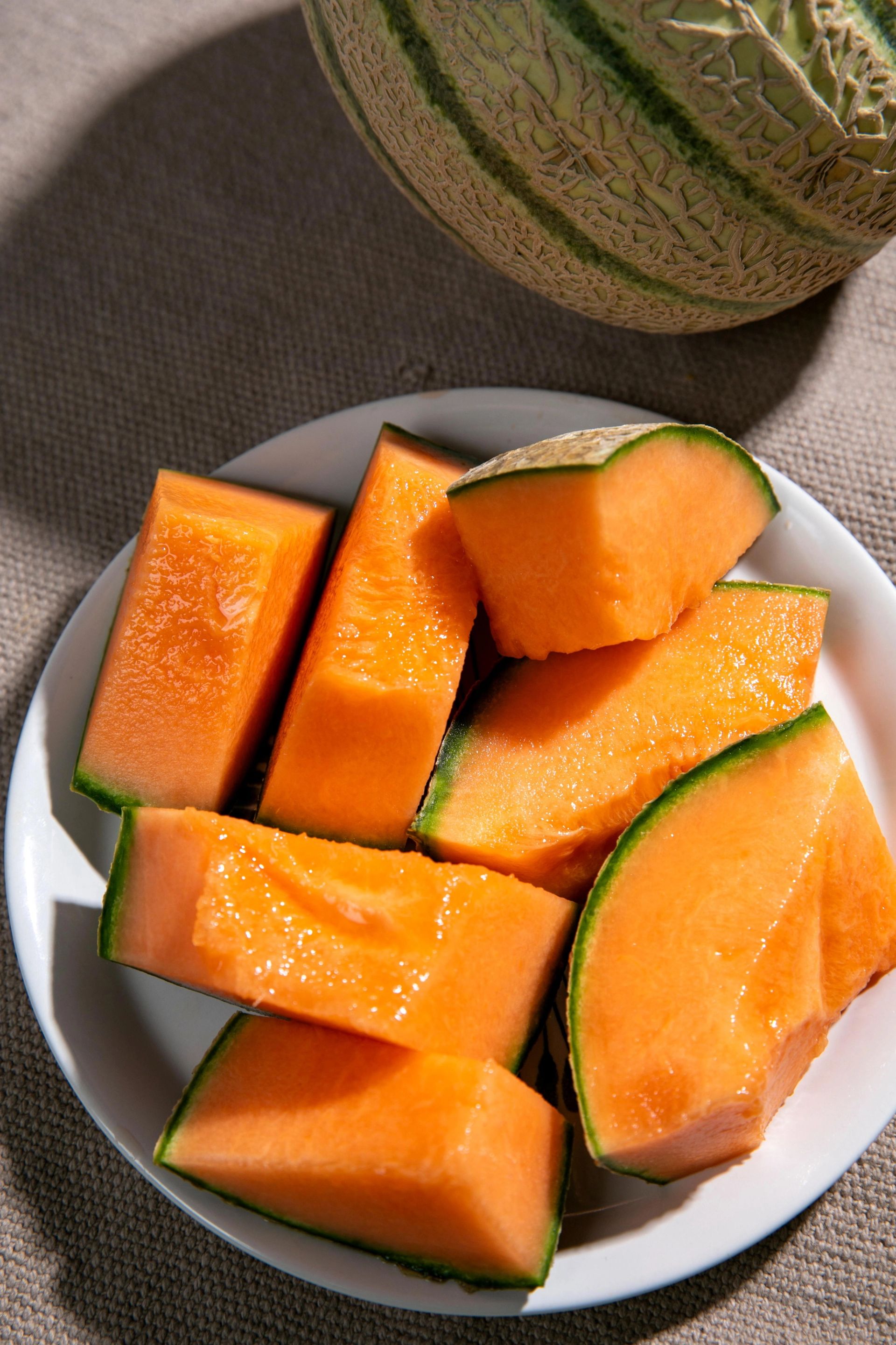 En hvit tallerken fylt med store biter av oransje cantaloupe‑melon med grønn kant, med en hel cantaloupe‑melon plassert bak tallerkenen