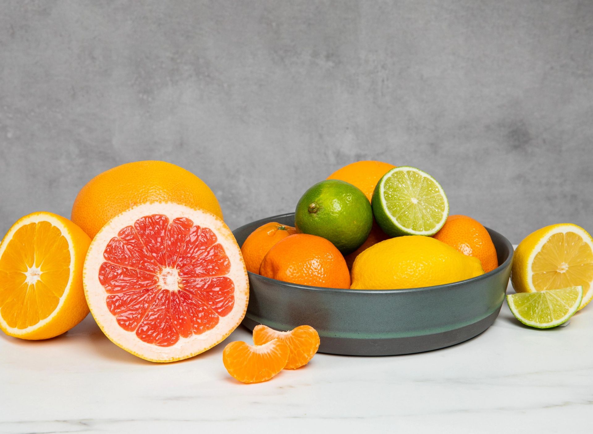 Et utvalg sitrusfrukter arrangert på en lys overflate, med grapefrukt delt i to, hele appelsiner, sitroner og lime i en mørk bolle, samt appelsinbåter og halve lime foran, mot en nøytral grå bakgrunn.