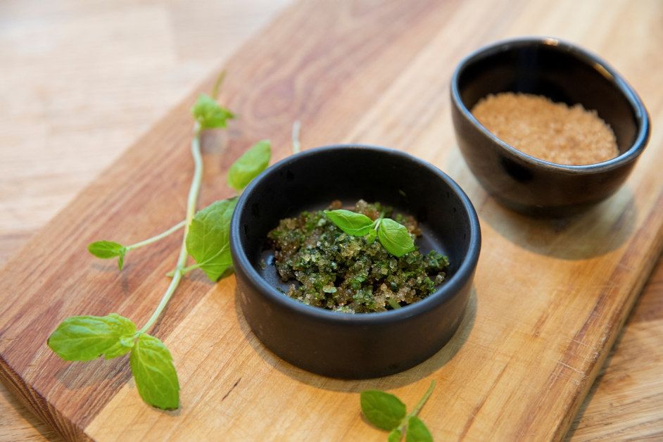To små, svarte keramikkskåler på et lyst trefat. Den fremste skålen inneholder grønn mintsukker, toppet med et friskt mintblad. I bakgrunnen, til høyre, er en skål med brunt sukker.