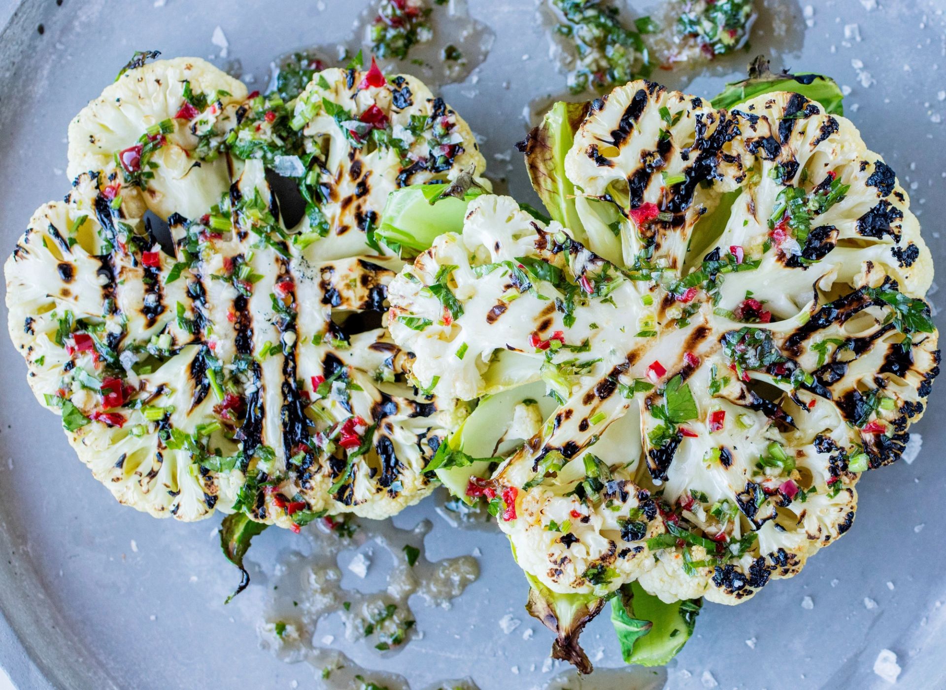 Grillet blomkål delt i to, med tydelige grillmerker, toppet med frisk urte- og chiliolje, hakkede urter og flaksalt