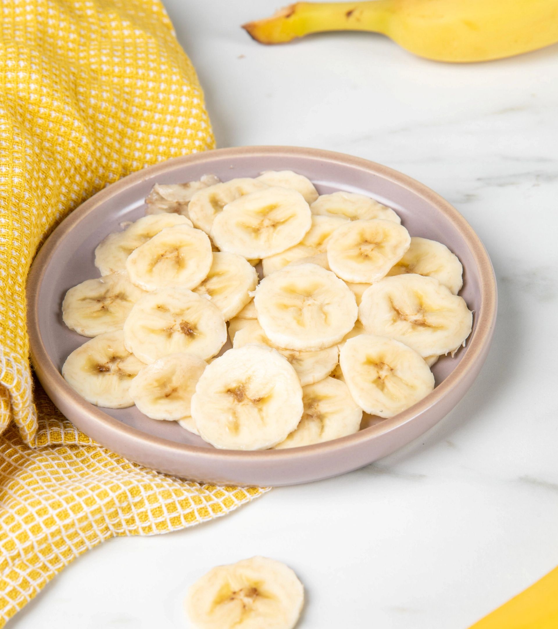 Banan i skiver ligger på en lys tallerken, pent fordelt i ett lag. Tallerkenen står på en lys benkeplate ved siden av et gult kjøkkenhåndkle, mens deler av hele bananer er synlige i bakgrunnen.