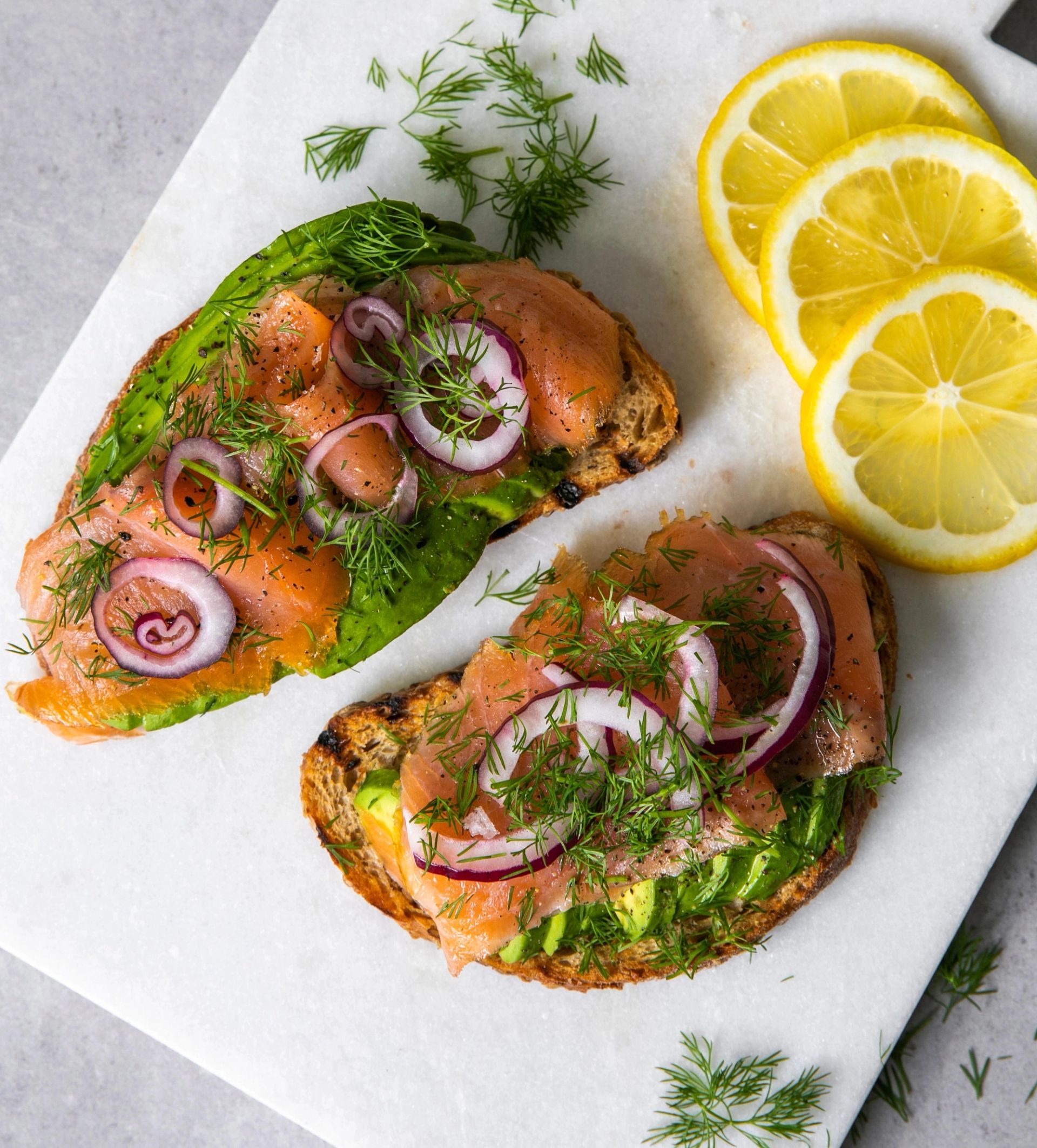 To skiver ristet brød med avokado, rødløk, røkt laks og frisk dill på et lyst brett, servert med tre skiver sitron ved siden av.