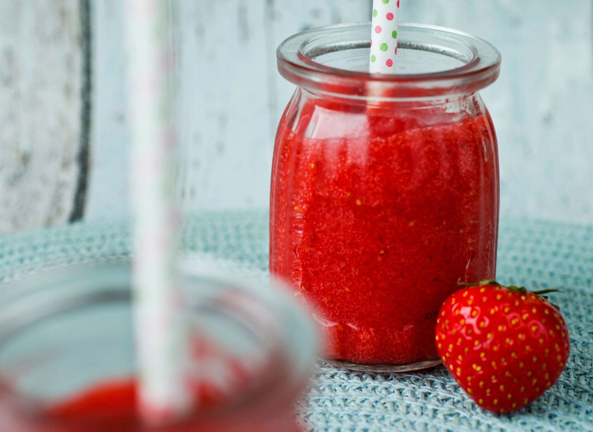 Et lite glass fylt med rød jordbærsmoothie står på et strikket underlag med et prikkete papirsugerør. Ved siden av glasset ligger et ferskt jordbær, mens et annet smoothieglass er uskarpt i forgrunnen.