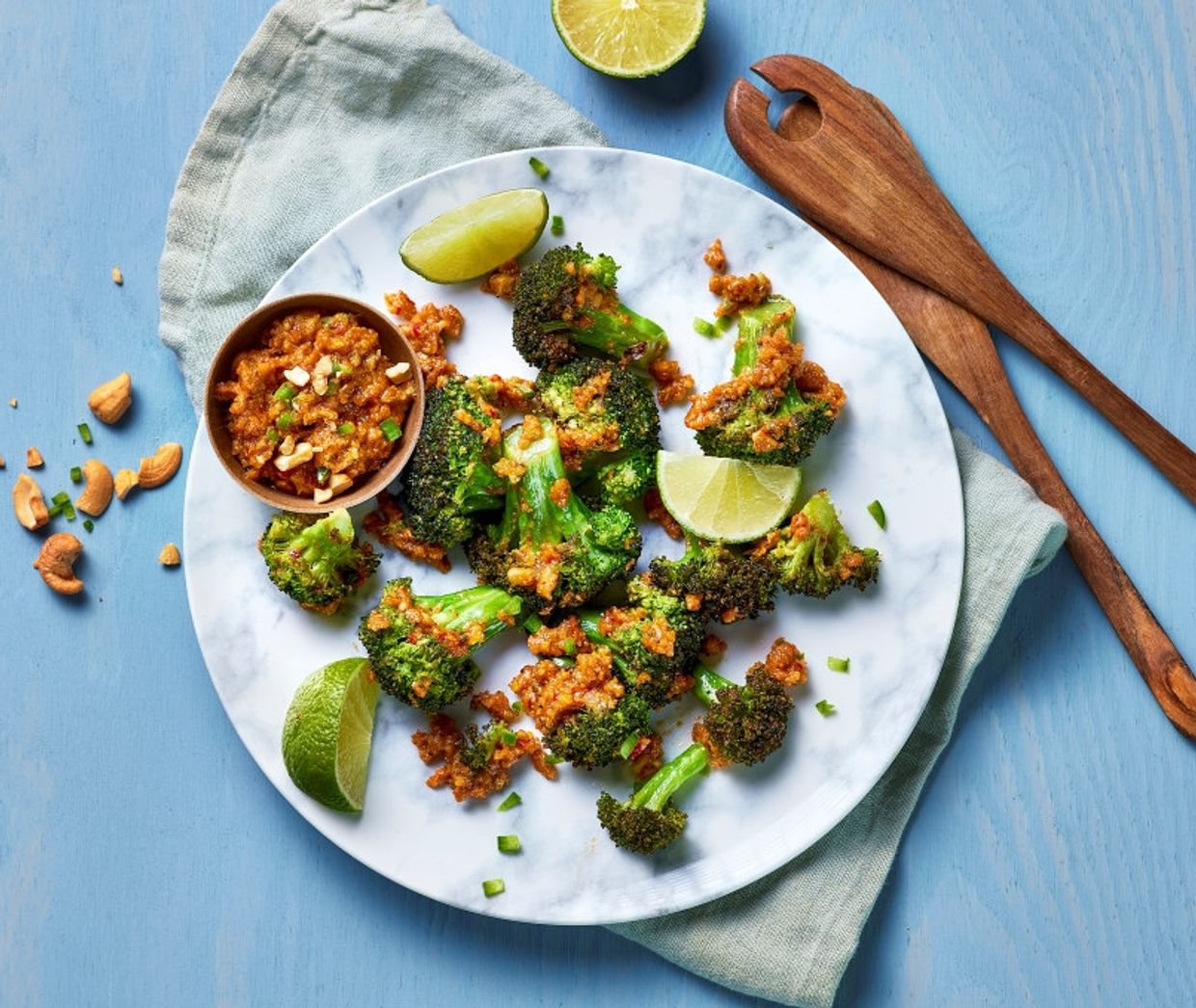 En tallerken med stekt brokkoli toppet med en gyllen topping, servert med limebåter. På tallerkenen står også en liten skål med saus. Rundt tallerkenen ligger noen cashewnøtter, en delt lime og finhakket grønt dryss. Tallerkenen er plassert på et lyst blått underlag med en linserviett og to tresleiver ved siden av.