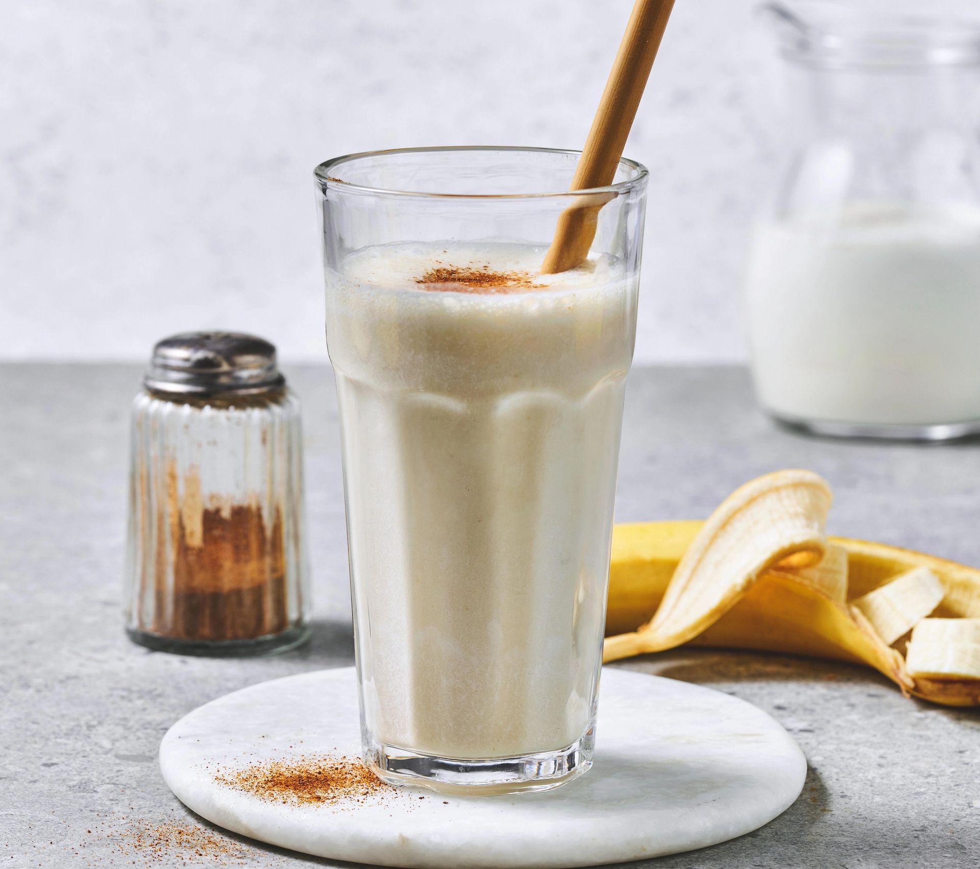 Et høyt glass med kremet banansmoothie står på en rund marmorfat med et sugerør i tre. Smoothien er toppet med et lett dryss kanel. Ved siden av glasset ligger en halvskrelt banan, og i bakgrunnen står en melkeflaske og en krydderbøsse på en lys grå benkeplate.