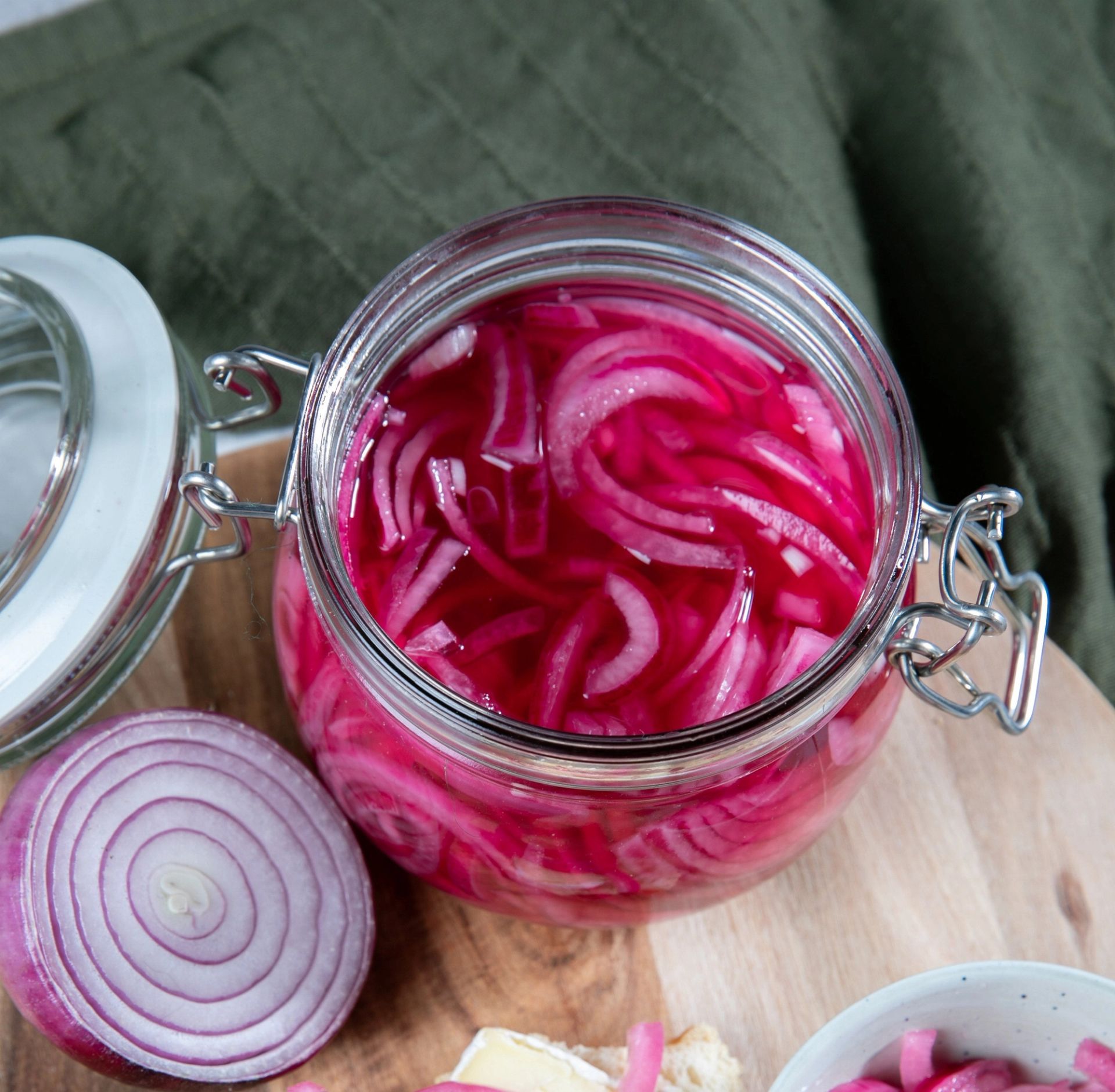 Et glass med syltet rødløk i skiver, fylt med klar rosa lake. Glasset står åpent på et trebrett, med en halv rå rødløk ved siden av.