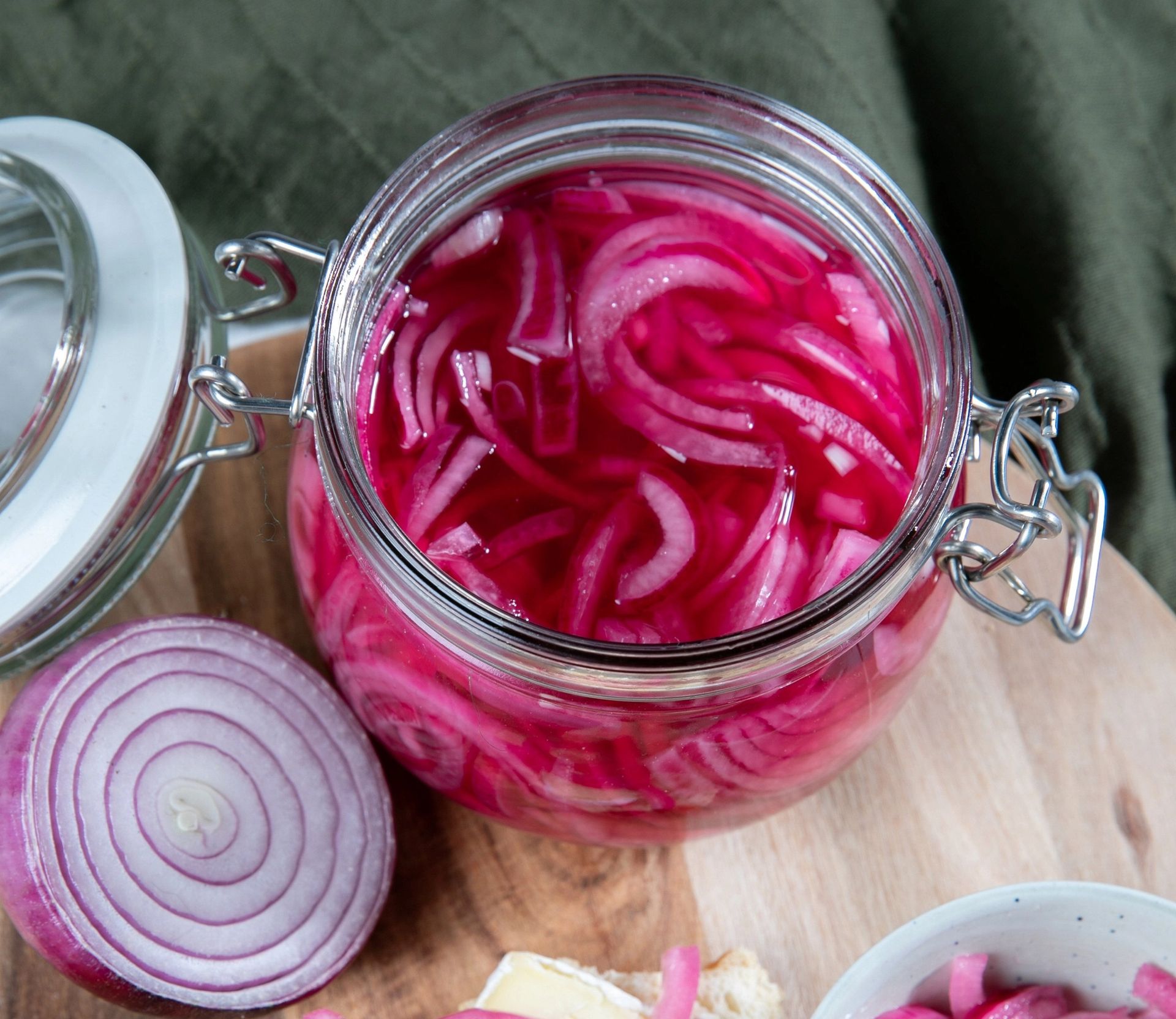Et glass med syltet rødløk i skiver, fylt med klar rosa lake. Glasset står åpent på et trebrett, med en halv rå rødløk ved siden av.