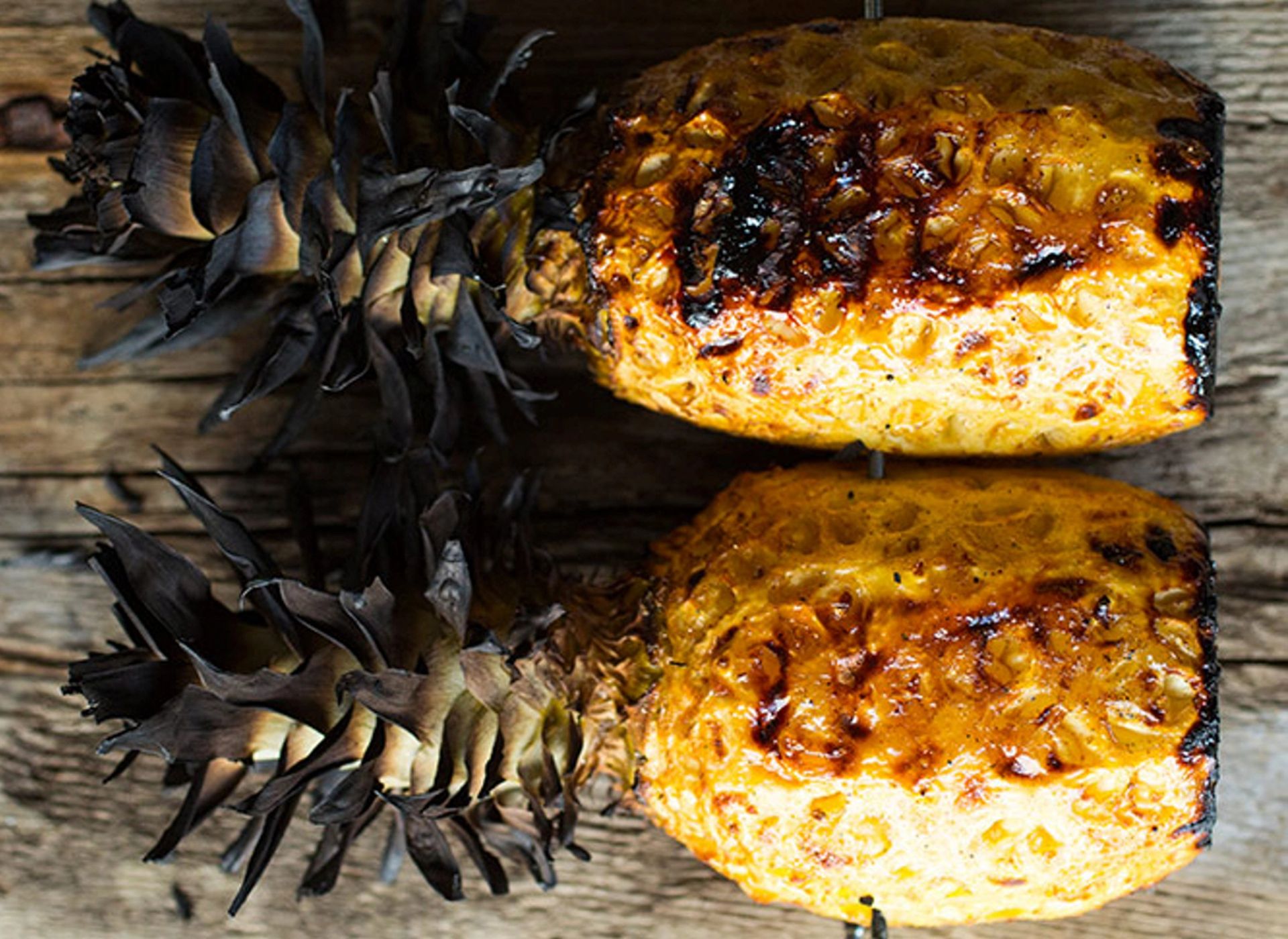 To hele ananaser ligger ved siden av hverandre på et rustikt treunderlag. Skallet er fjernet, og overflaten er grillet slik at den har en gyllen farge med mørkere karamelliserte og lett svidde partier. Toppen med de tørre, mørke bladene er fortsatt festet på begge ananasene.