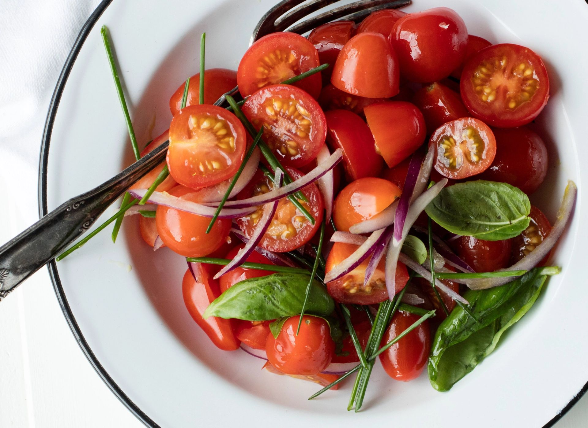En frisk tomatsalat med halverte cherrytomater, tynne skiver rødløk, grønn basilikum og gressløk, servert på en hvit tallerken med en skje ved siden av.
