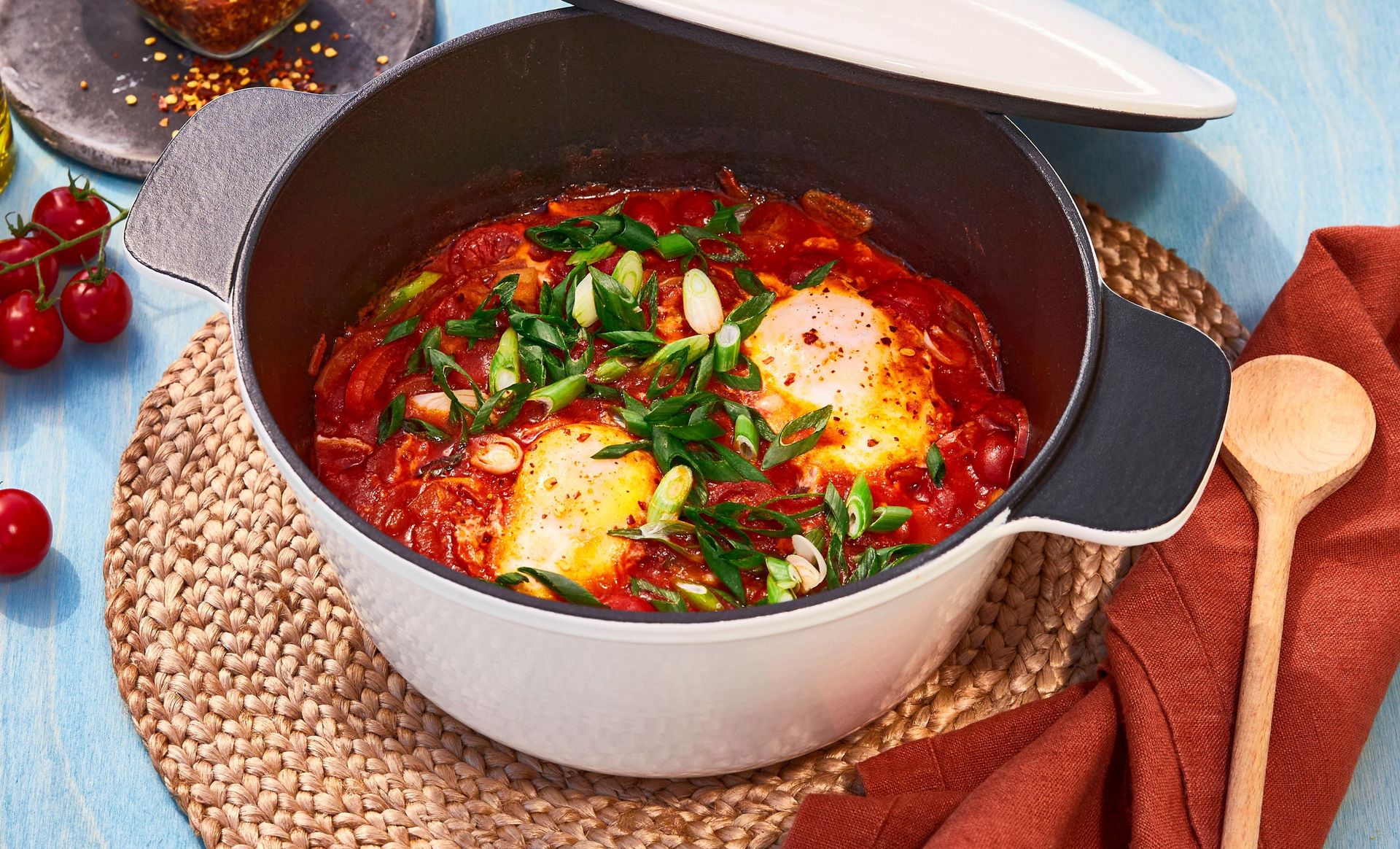 Shakshuka servert i en rund, hvit gryte med svart innside og halvveis åpent lokk. To posjerte egg ligger i den fyldige, røde tomatsausen, rikelig toppet med snittet vårløk og et dryss krydder.